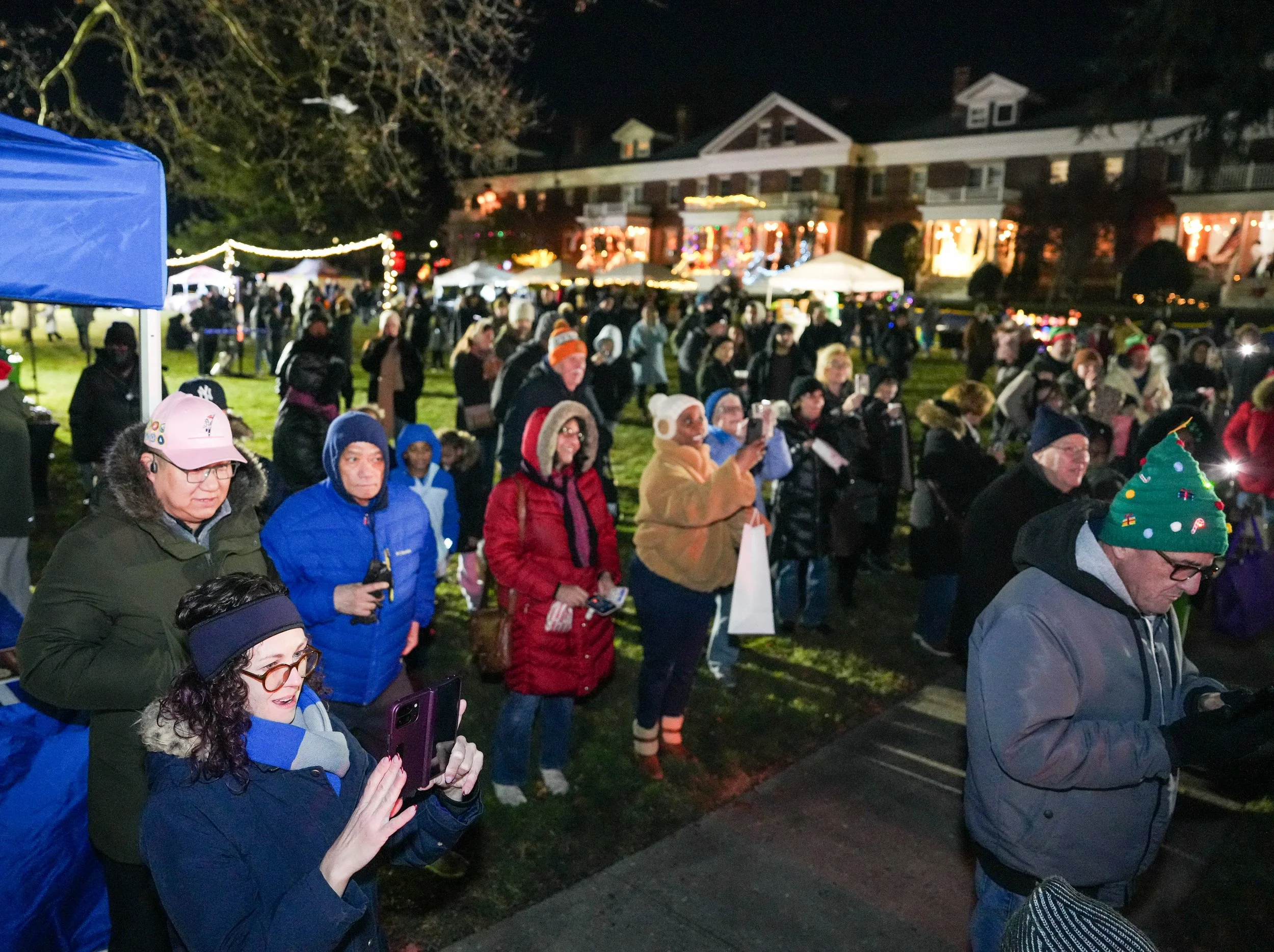 A large crowd of people gathered outdoors at night, many wearing warm clothing and holiday accessories, in front of a row of houses decorated with Christmas lights and ornaments, likely for a holiday event or celebration.
