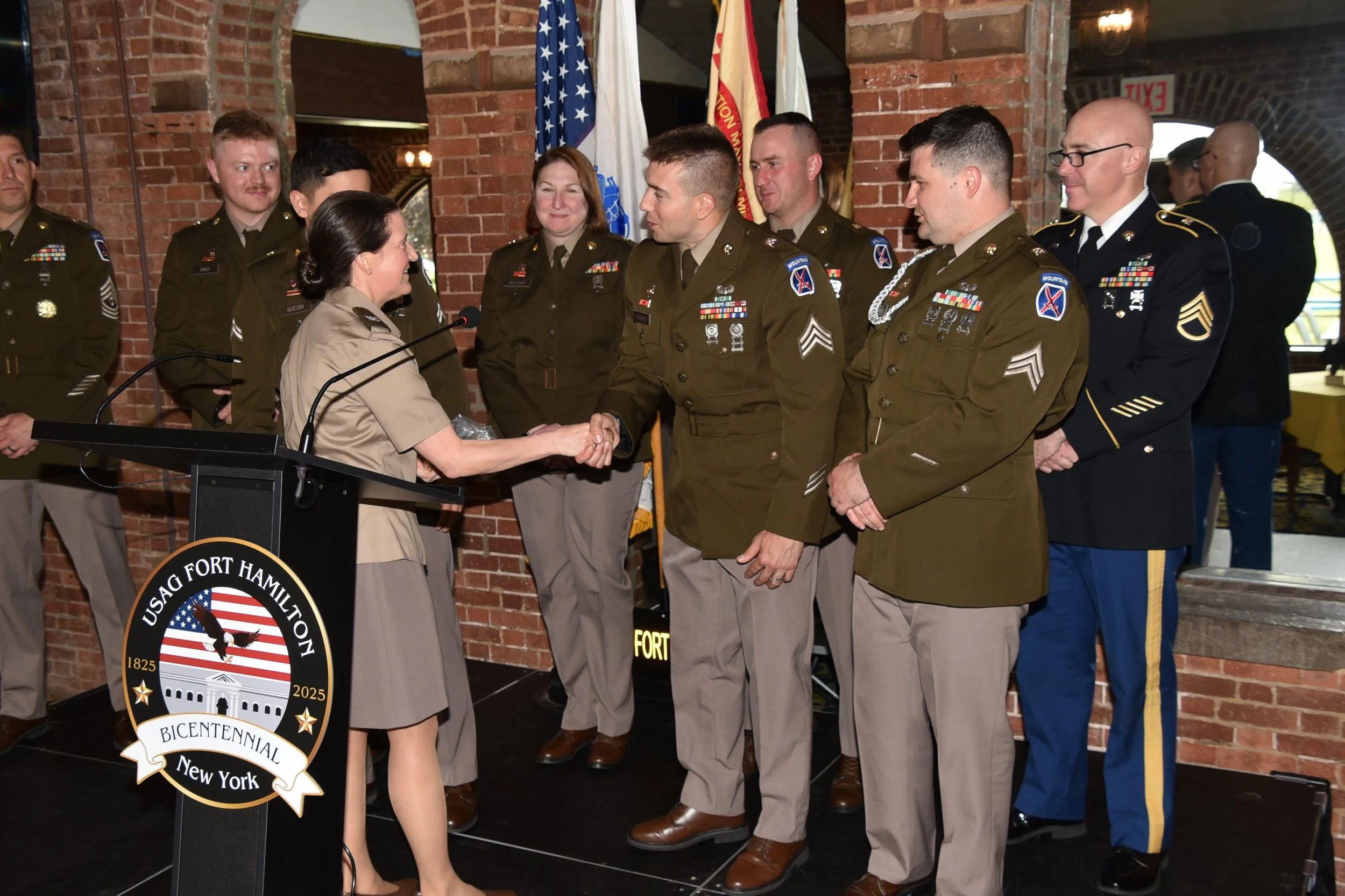 A group of military personnel, both men and women, are standing on a stage with brick walls and flags in the background. Two women are shaking hands in the center, with others watching and smiling. The scene appears to be a formal ceremony or award presentation at Fort Hamilton, New York.