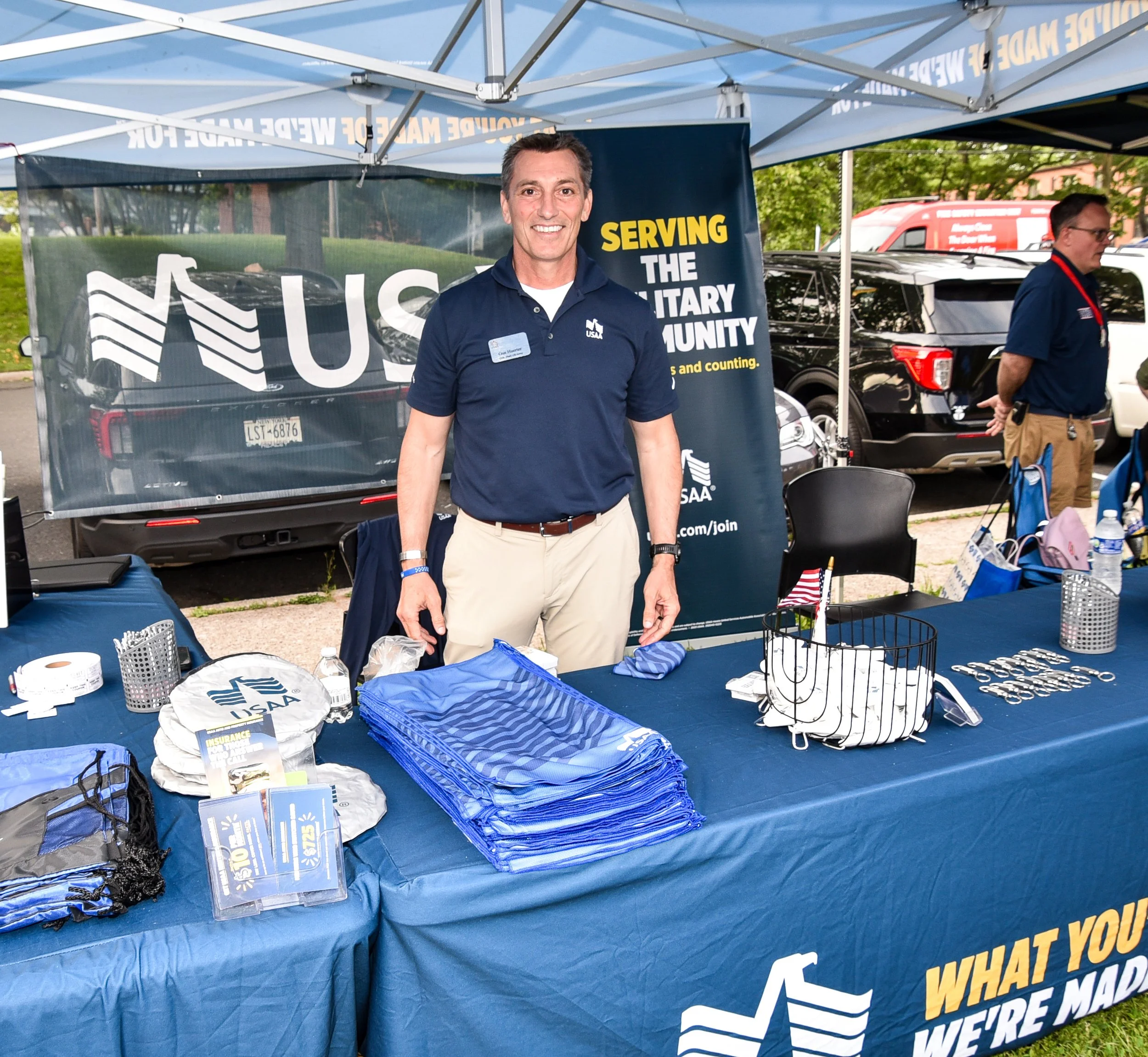 A man wearing a navy polo shirt with a name tag standing at an outdoor booth with Canvases USA branding. The booth has a blue tablecloth, promotional items, and a large banner behind him that says "Serving the Military Community." There are other people, vehicles, and trees in the background.