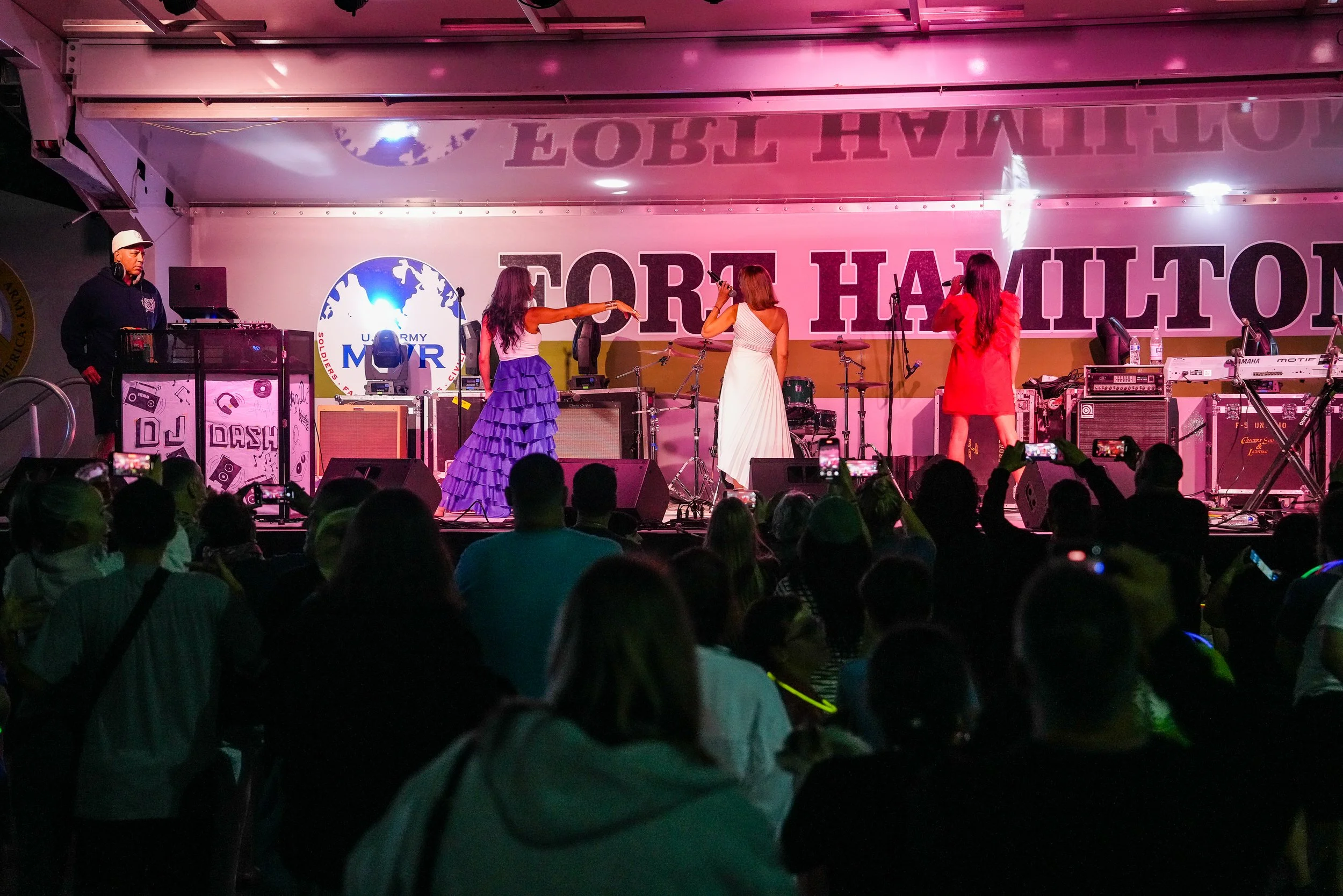 A stage with three women singing into microphones and a DJ on the left. The backdrop has the words 'FORT HAMILTON' and a logo with a globe and the letters 'U.S. ARMY MWR'. Audience members are visible in the foreground.