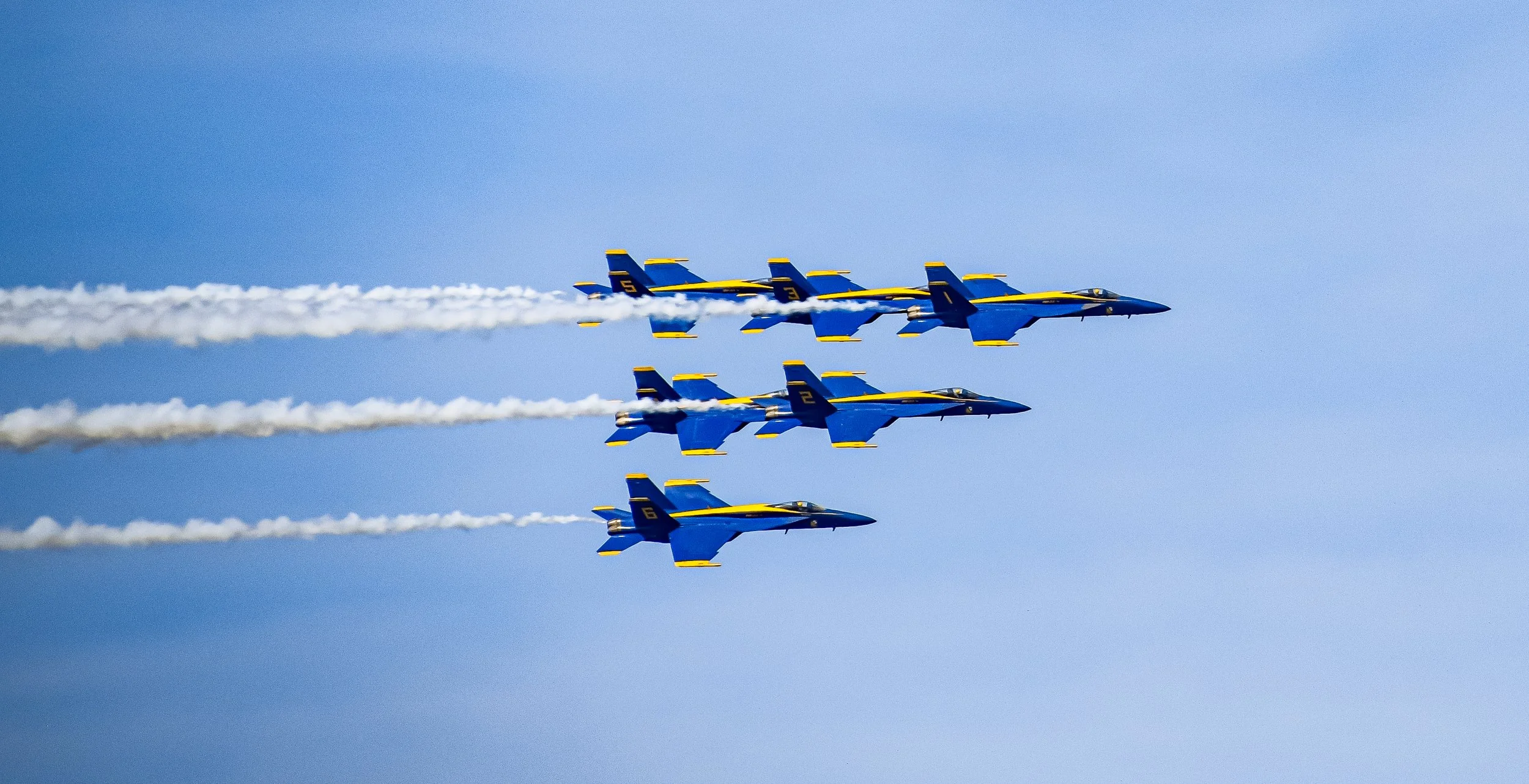 A lineup of six blue fighter jets flying in formation through a clear blue sky, emitting white smoke trails.
