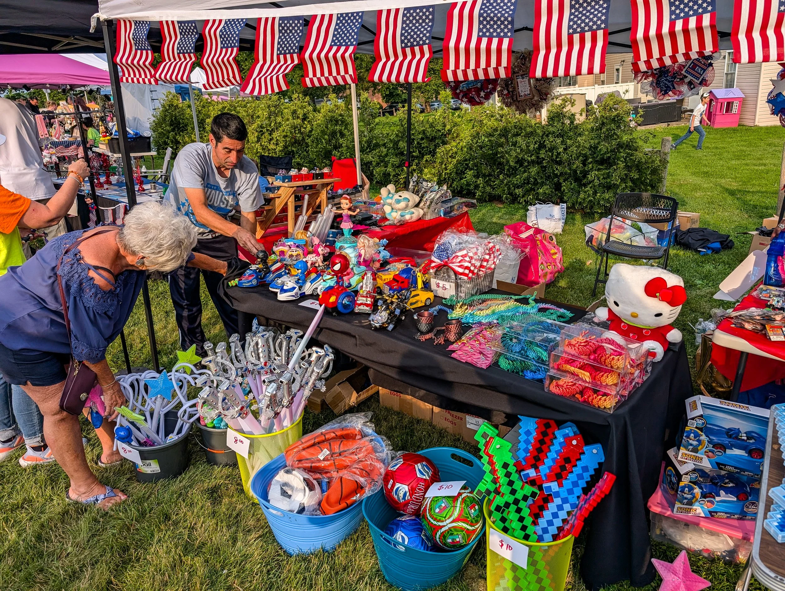 A vendor stall at an outdoor market decorated with American flags, displaying various toys and jewelry. People browsing and a girl running in the background, with a grassy area and houses in the distance.