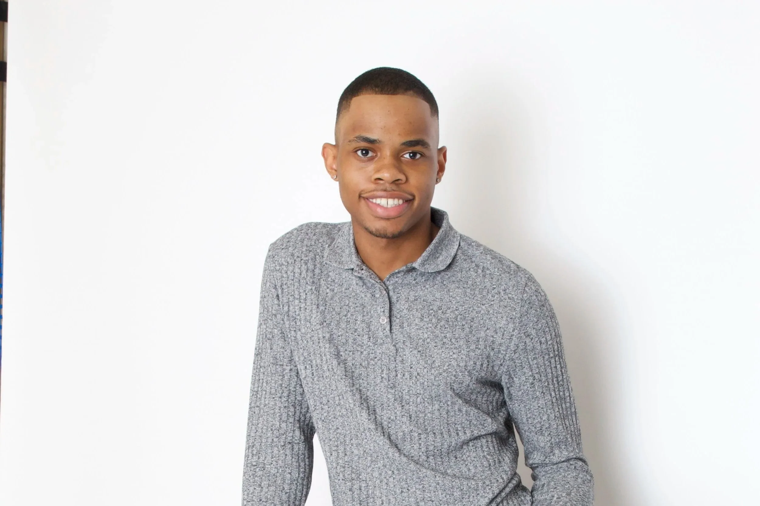 Young man with short hair, wearing a gray patterned button-up shirt, smiling, sitting against a plain white background.