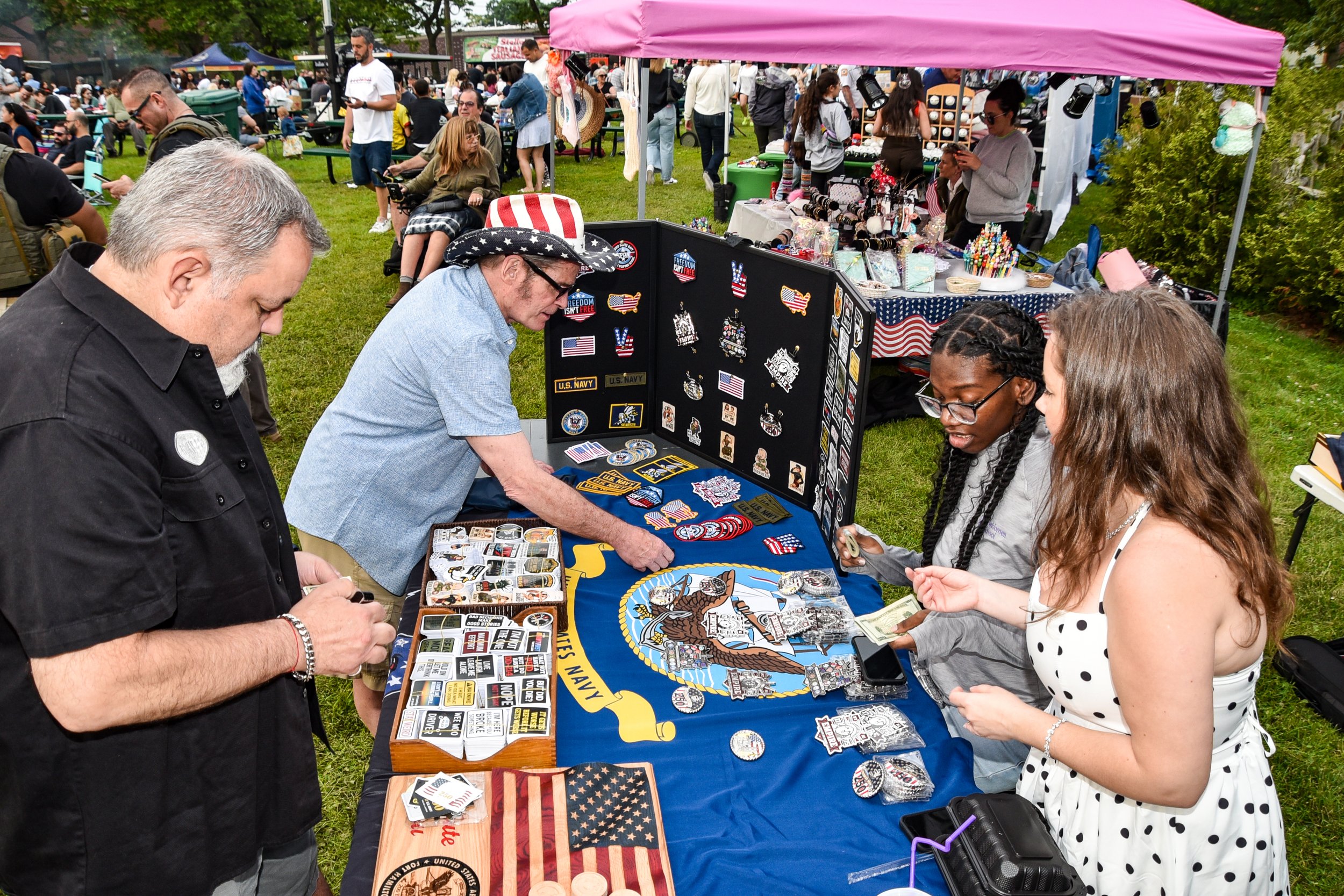 People browsing patriotic patches, pins, and merchandise at an outdoor market stall during a festival or gathering, with a crowd and vendor tents in the background.