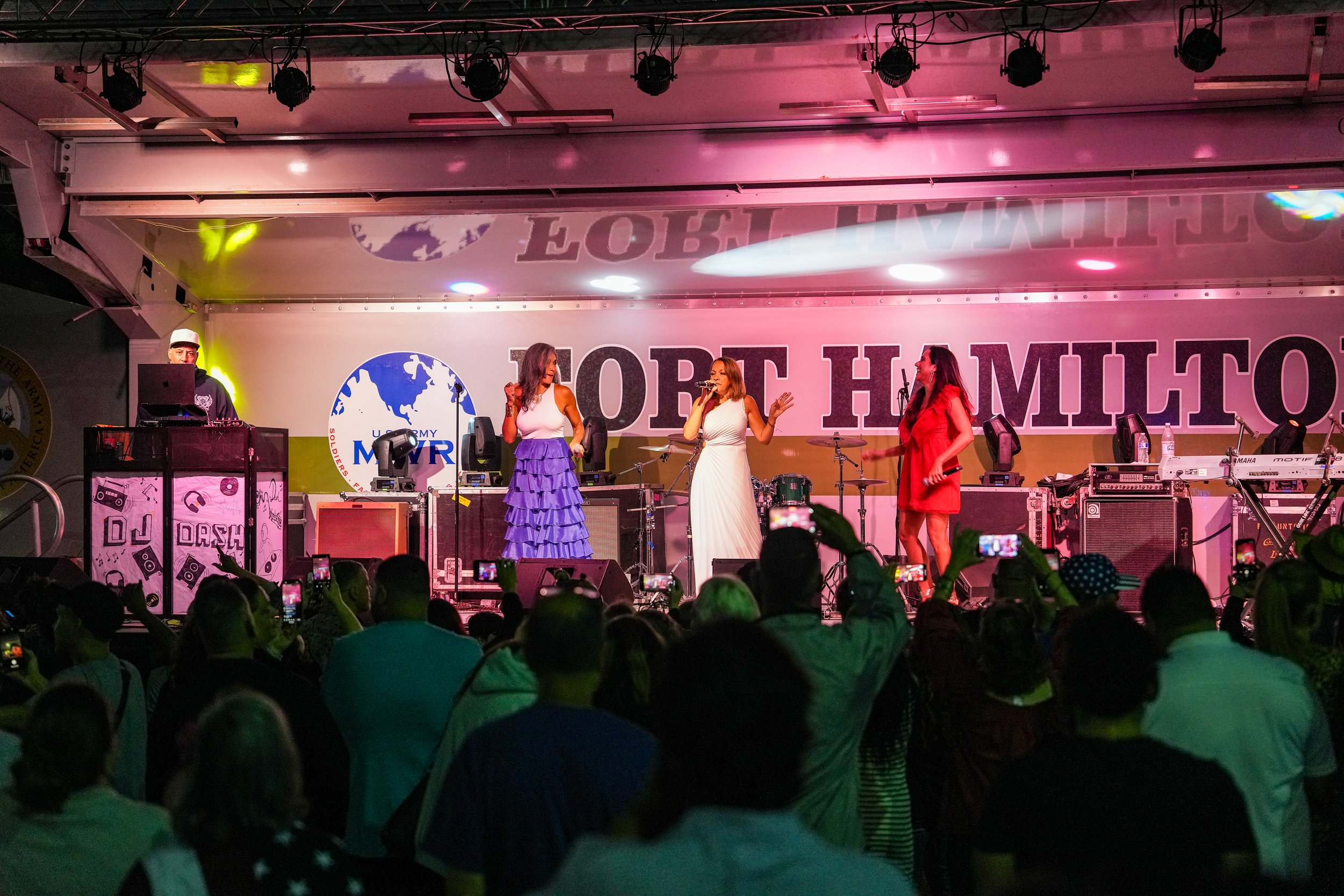 Women singing on stage at an event with a crowd watching, with a large banner behind them reading "TOLE HAMITO." A DJ is on the left, and there are musical instruments and sound equipment around them.