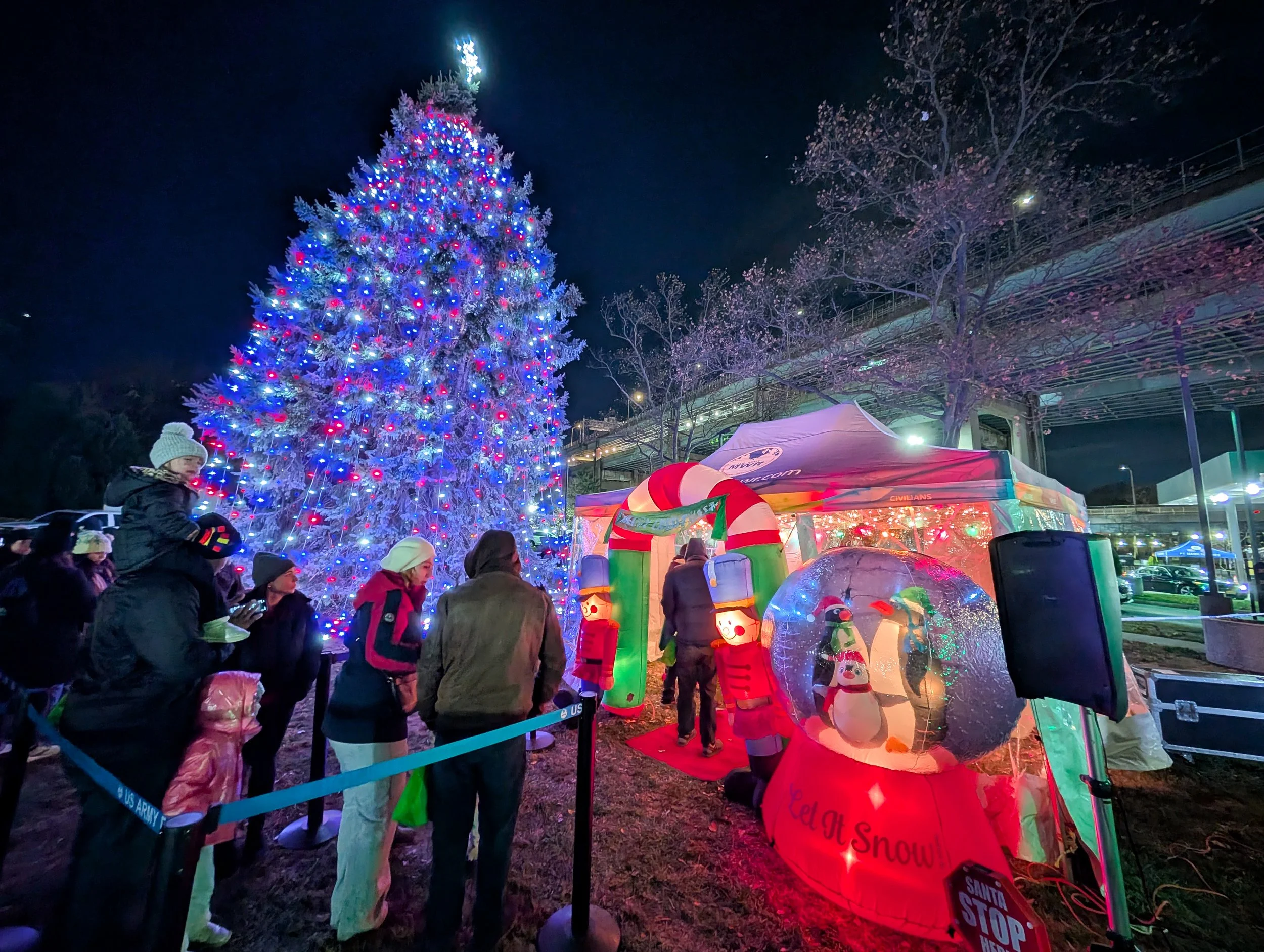 People standing in line outside at night near a large, decorated Christmas tree with colorful lights. There are Christmas-themed inflatable decorations and a small tent.