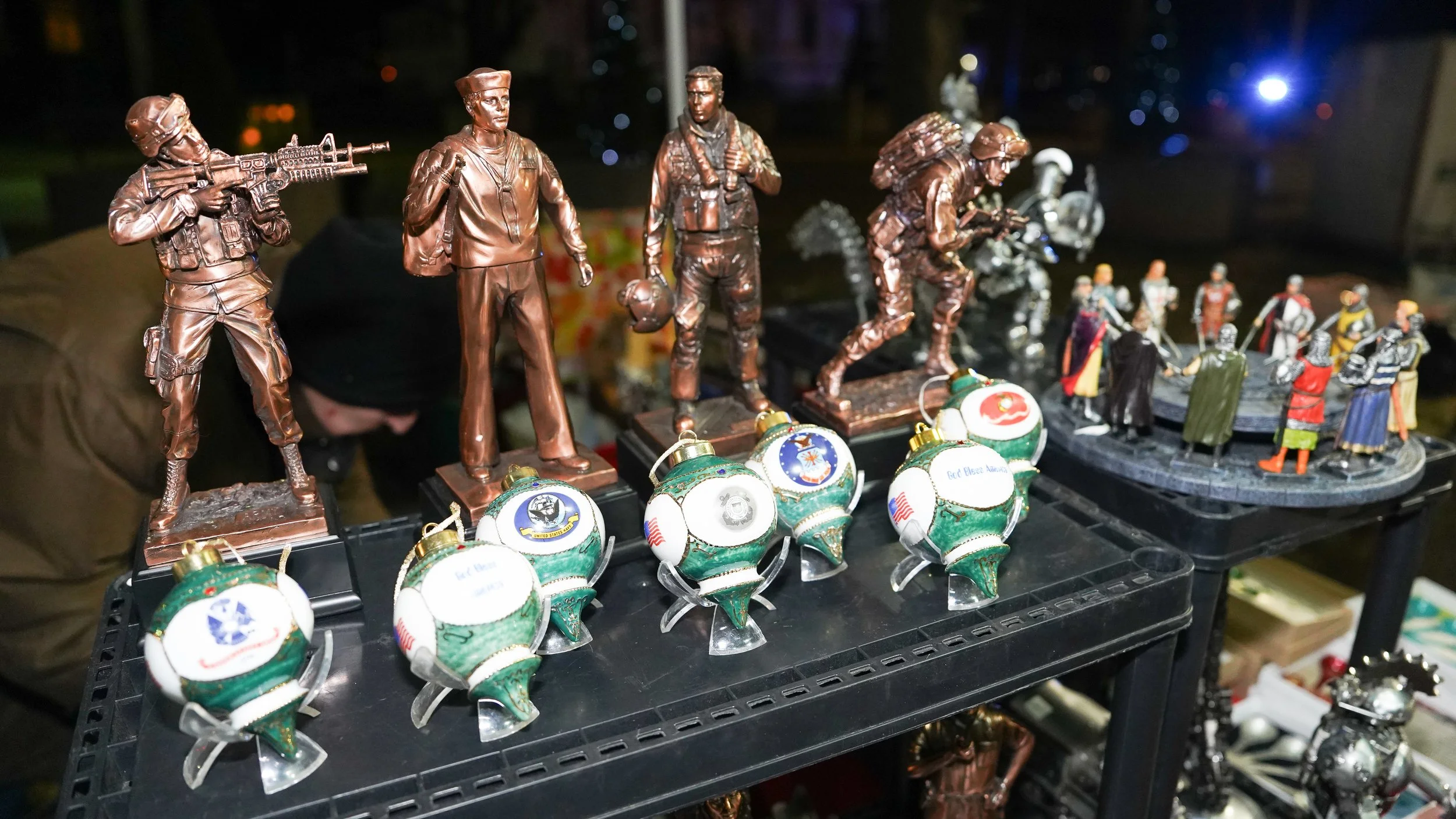 Display of bronze figurines representing soldiers and civilians, with some holding guns and others carrying bags, alongside decorative glass ornaments.