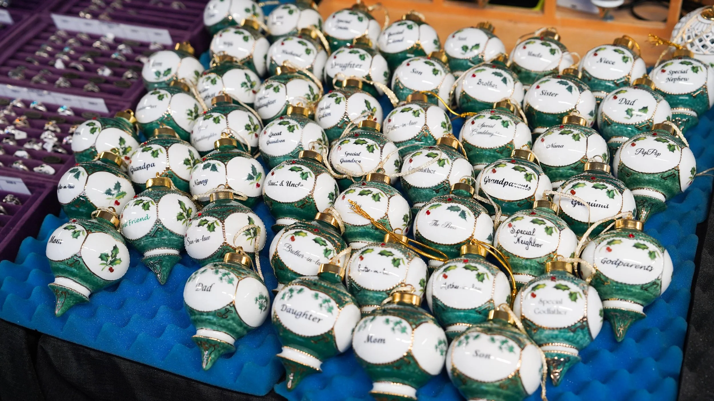 Christmas ornaments with green and white design, decorated with holly and red berries, inscribed with family relationship labels on a display at a market.
