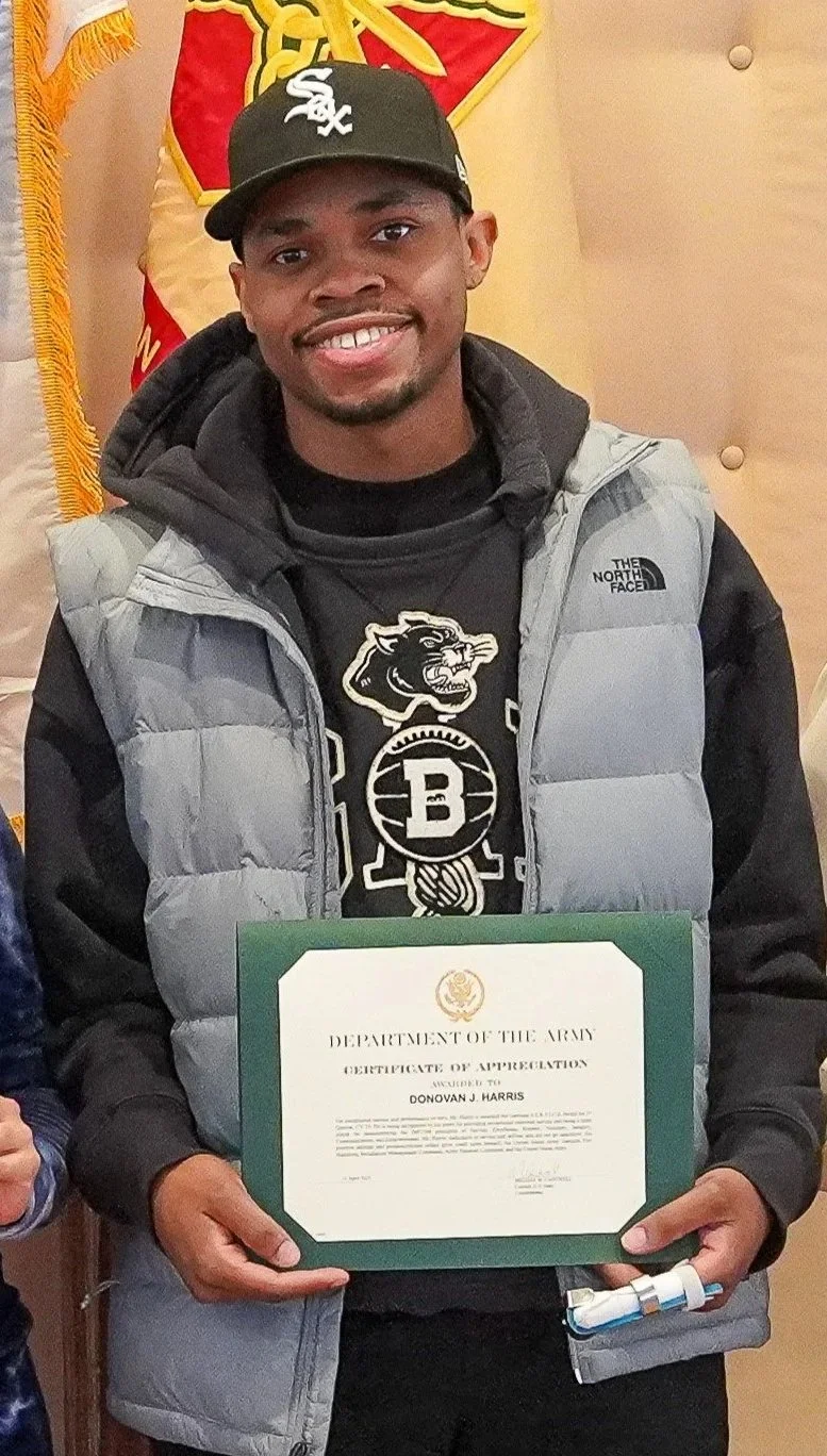 Young man smiling and holding a certificate of appreciation from the Department of the Army, wearing a black baseball cap and a gray puffer vest, with a black hoodie featuring the Brooklyn Nets logo, and standing in front of a flag and a beige background.