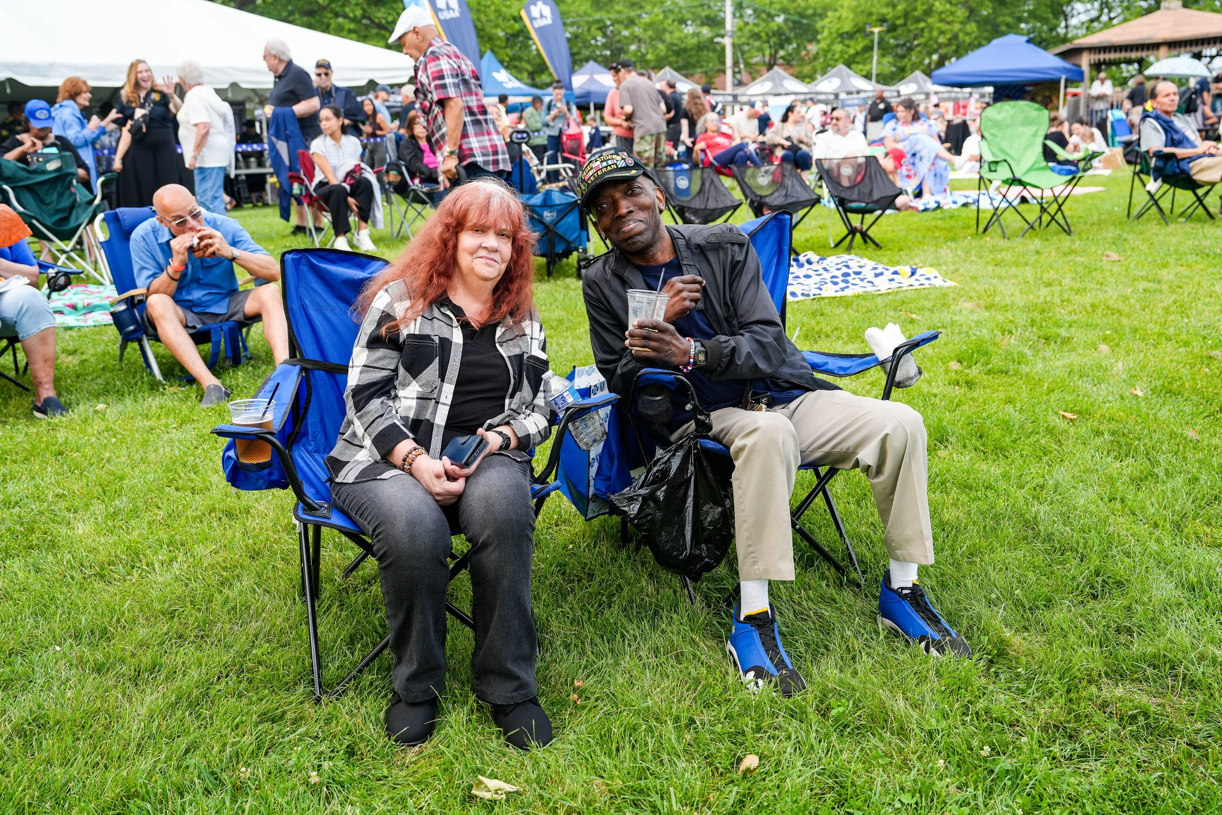 People gathered outdoors at a festival, with some sitting on lawn chairs, including a woman with red hair and a plaid shirt and a man with a cap and dark jacket, holding a drink. In the background, there are tents, flags, and more attendees sitting on the grass.