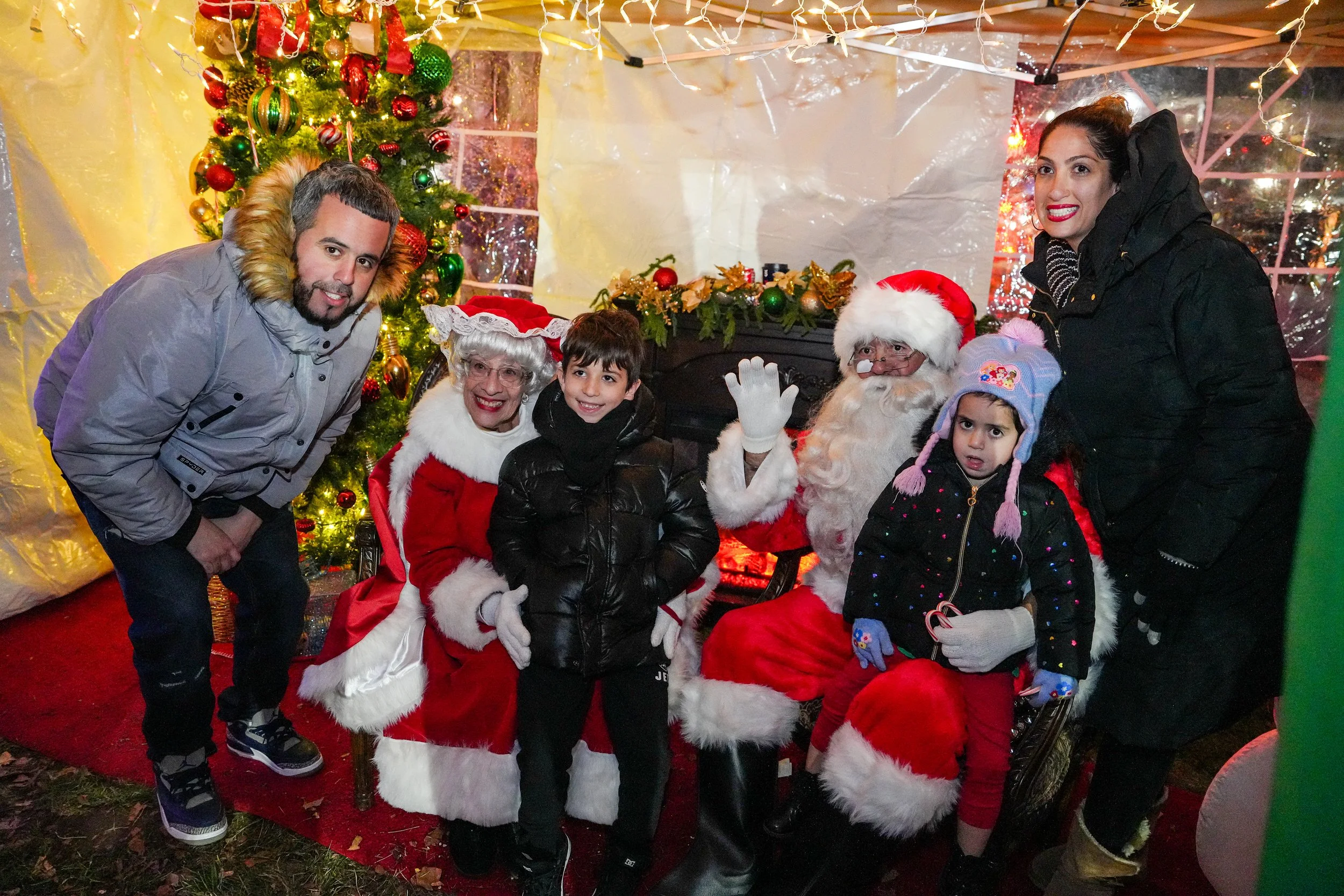 People posing with Santa Claus at a Christmas event. There is a Christmas tree decorated with red, green, and gold ornaments, and holiday decorations in the background. The group includes a man in a gray jacket, a woman dressed as Mrs. Claus, a young boy in black, another woman standing, and two children, one girl with a pink hat and another girl with a blue hat.