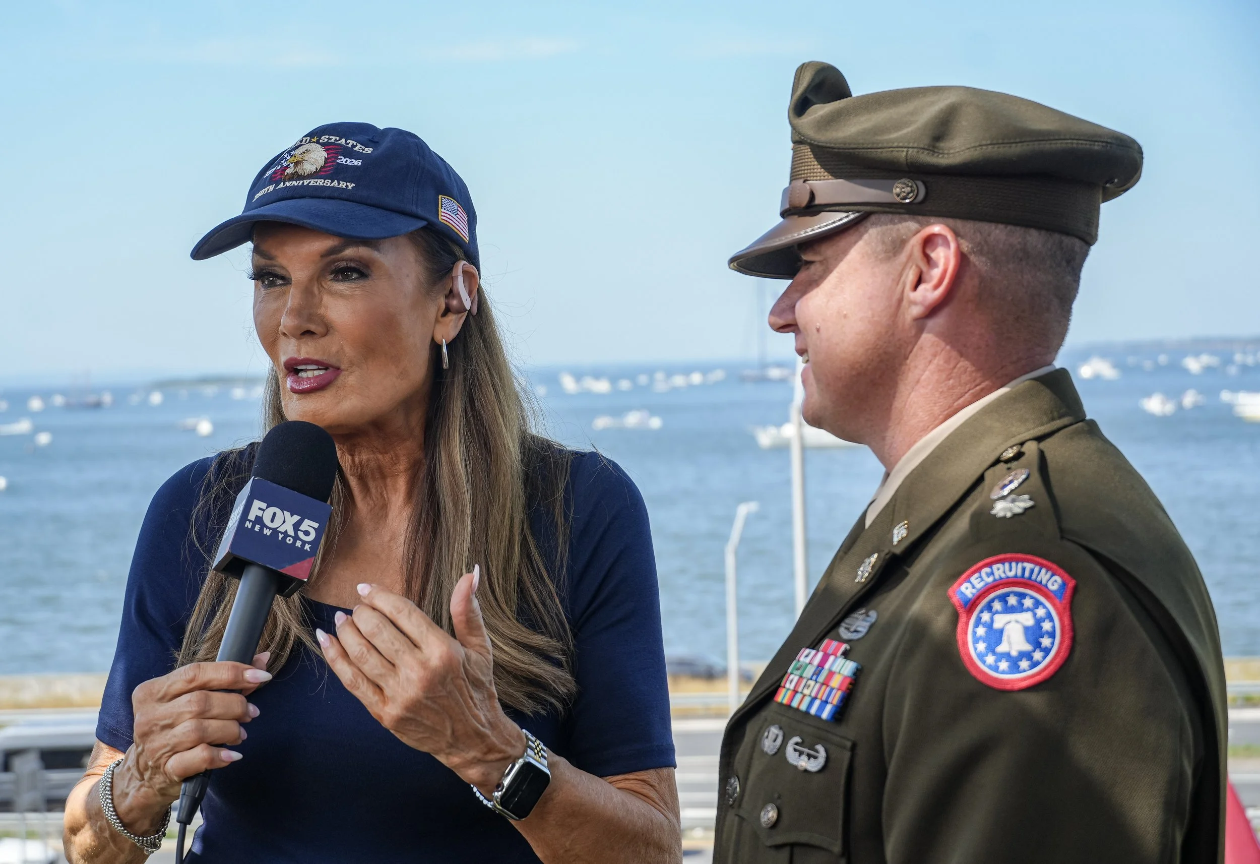 A woman with long brown hair and a man in a military uniform having a conversation outdoors near a body of water with boats in the background. The woman is holding a microphone labeled 'FOX 5 NEW YORK' and wearing a blue cap with American flag patches. The man is wearing a green military uniform with several medals and a patch that reads 'RECRUITING'.