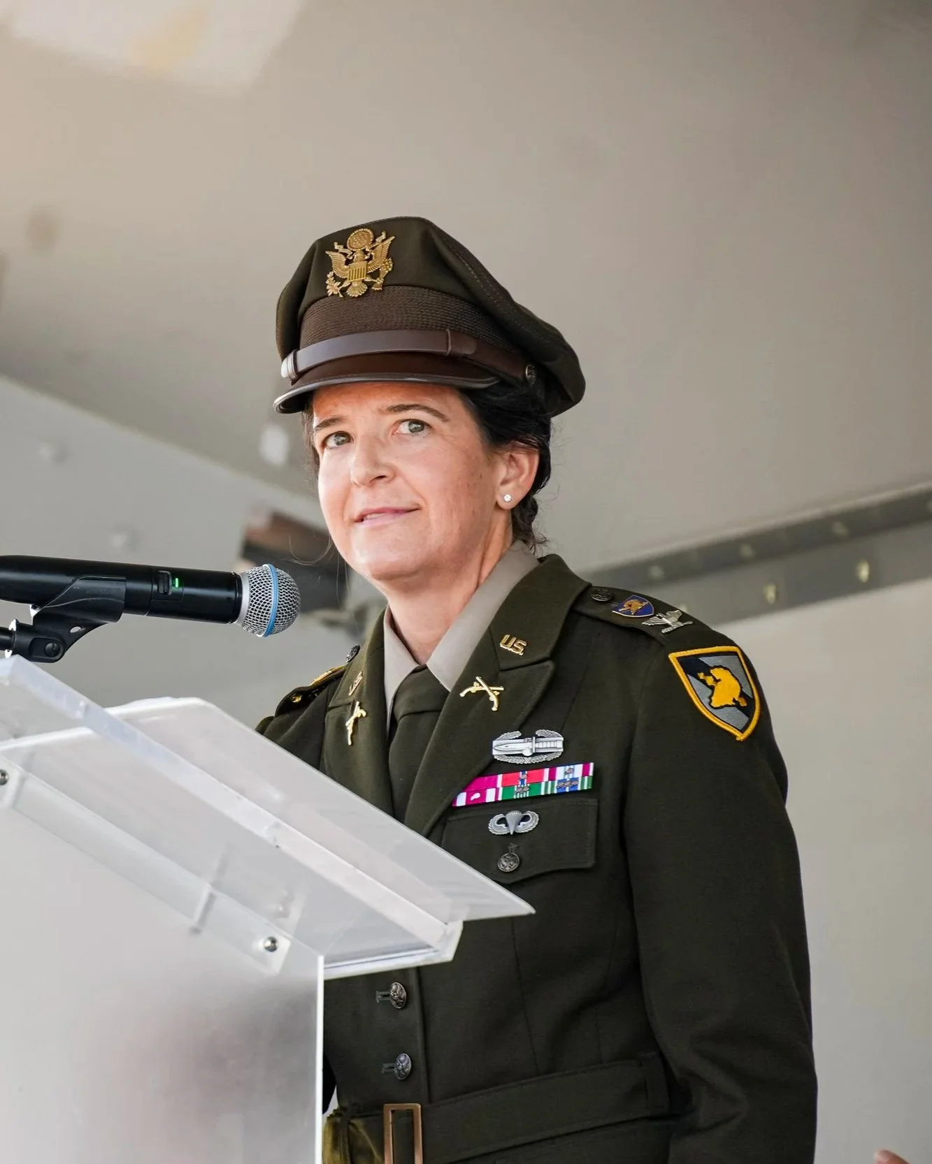 A woman in military uniform wearing a U.S. Army dress hat, standing at a podium, speaking into a microphone during a formal event.