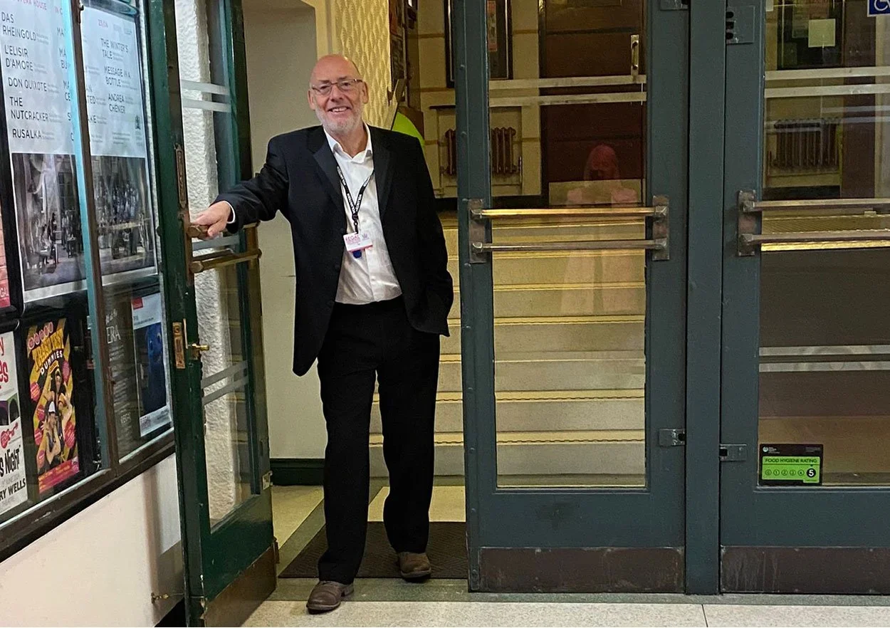 A man in a black suit and glasses standing at the entrance of a building, holding the door open and smiling.