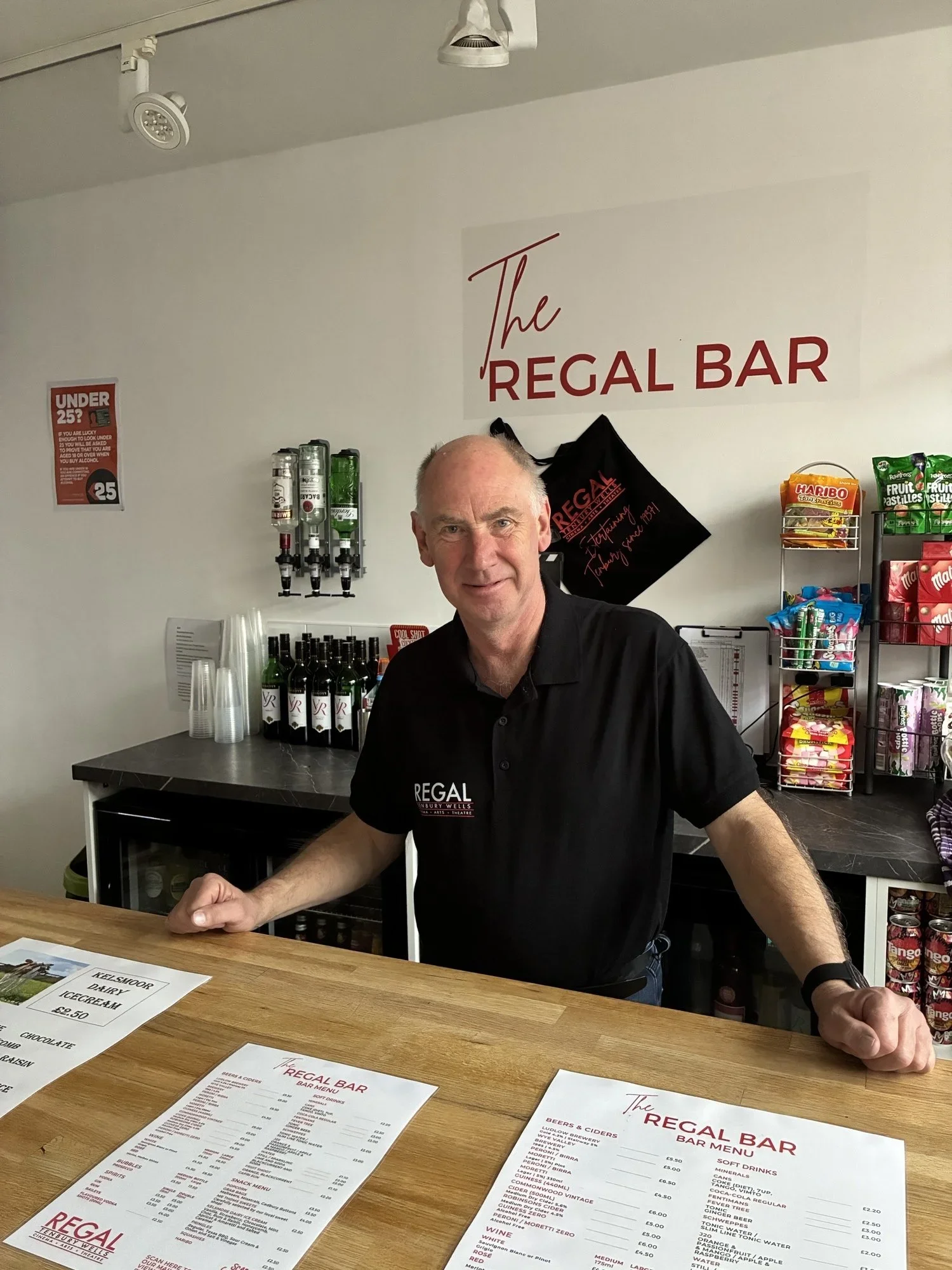A man behind a bar counter with menus in front, bar equipment behind him, and signs on the wall, including 'The Regal Bar'.