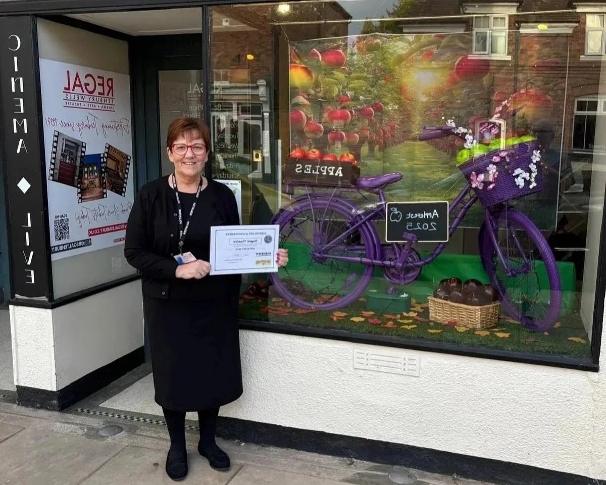 A woman standing in front of a store window holding a certificate. The store window displays a purple bicycle with a basket of flowers and apples, with a sign indicating 'Apple's 2025'.