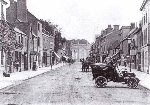 Black and white photo of a town street with buildings on both sides and a vintage car parked in the foreground.