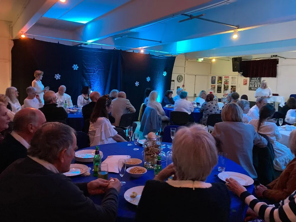 A group of people seated at round tables in a dimly lit room decorated with snowflakes on a black curtain backdrop, attending a formal event with food and drinks.