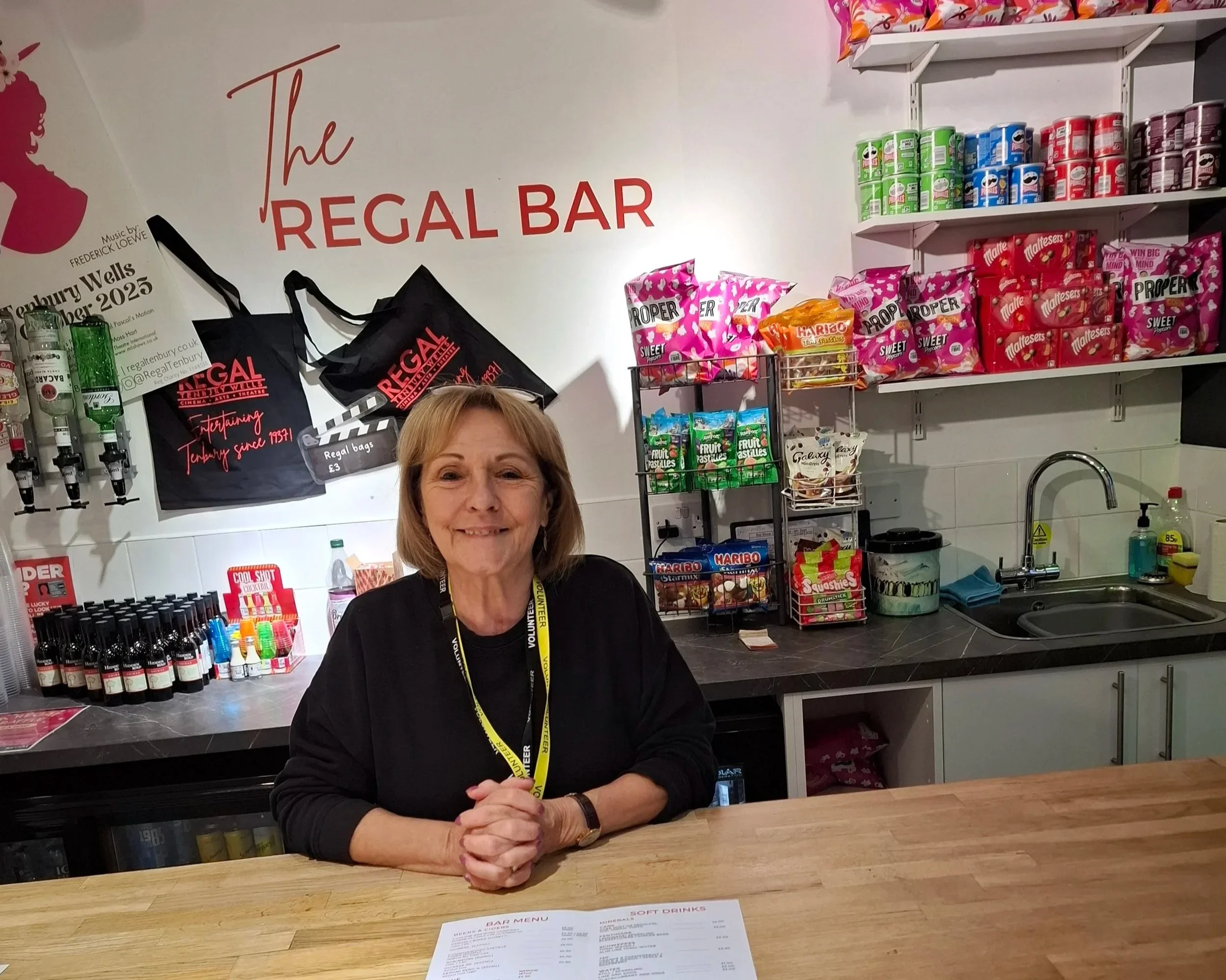 A woman with light brown hair sitting at a bar counter smiling at the camera. Behind her are shelves filled with canned drinks, bags of snacks, and other bar supplies. The wall has a large sign that reads 'The Regal Bar' with some smaller promotional materials and black bags hanging on the wall.