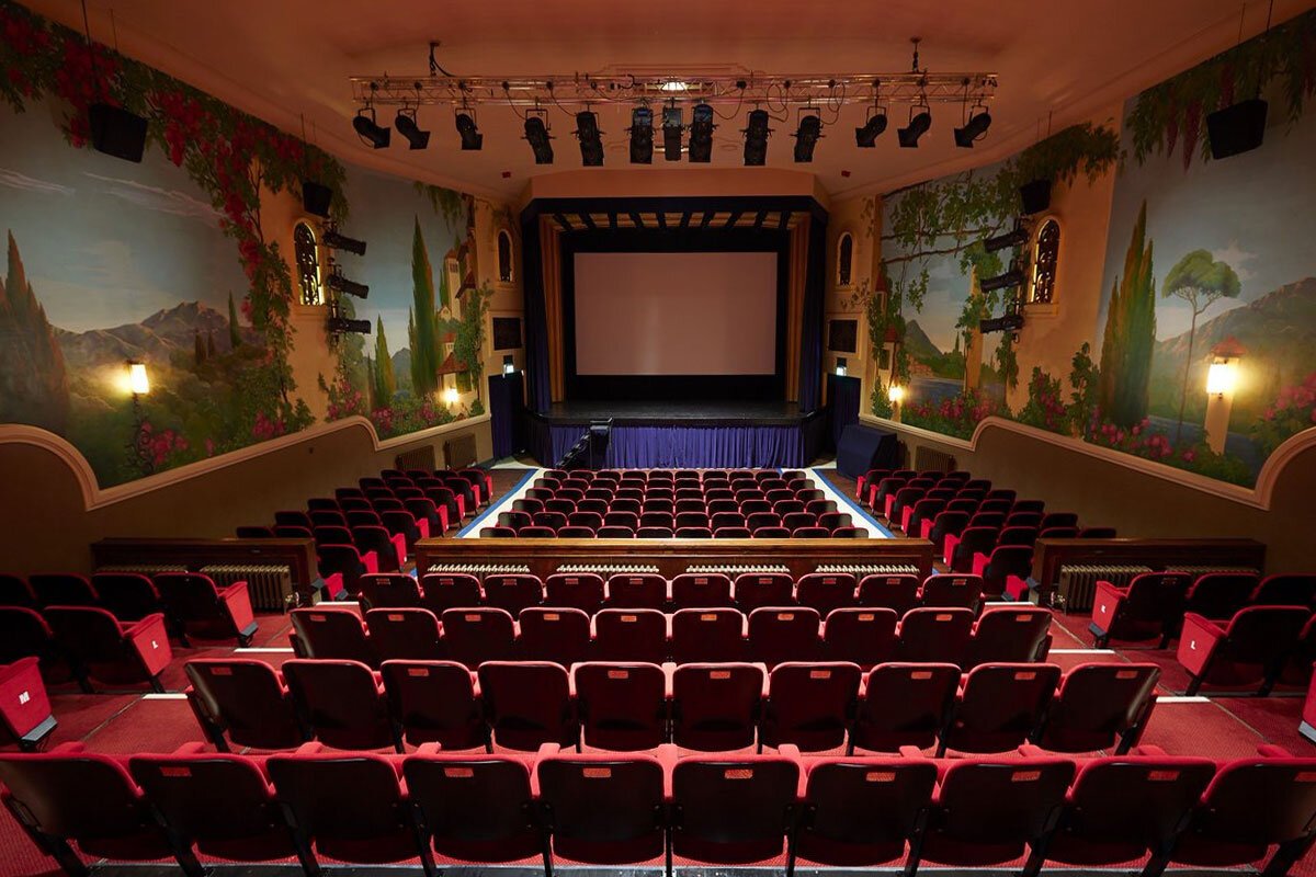 Empty theater with red seats facing a stage with a large white screen. Wall murals depict a scenic landscape with mountains, trees, and flowers.