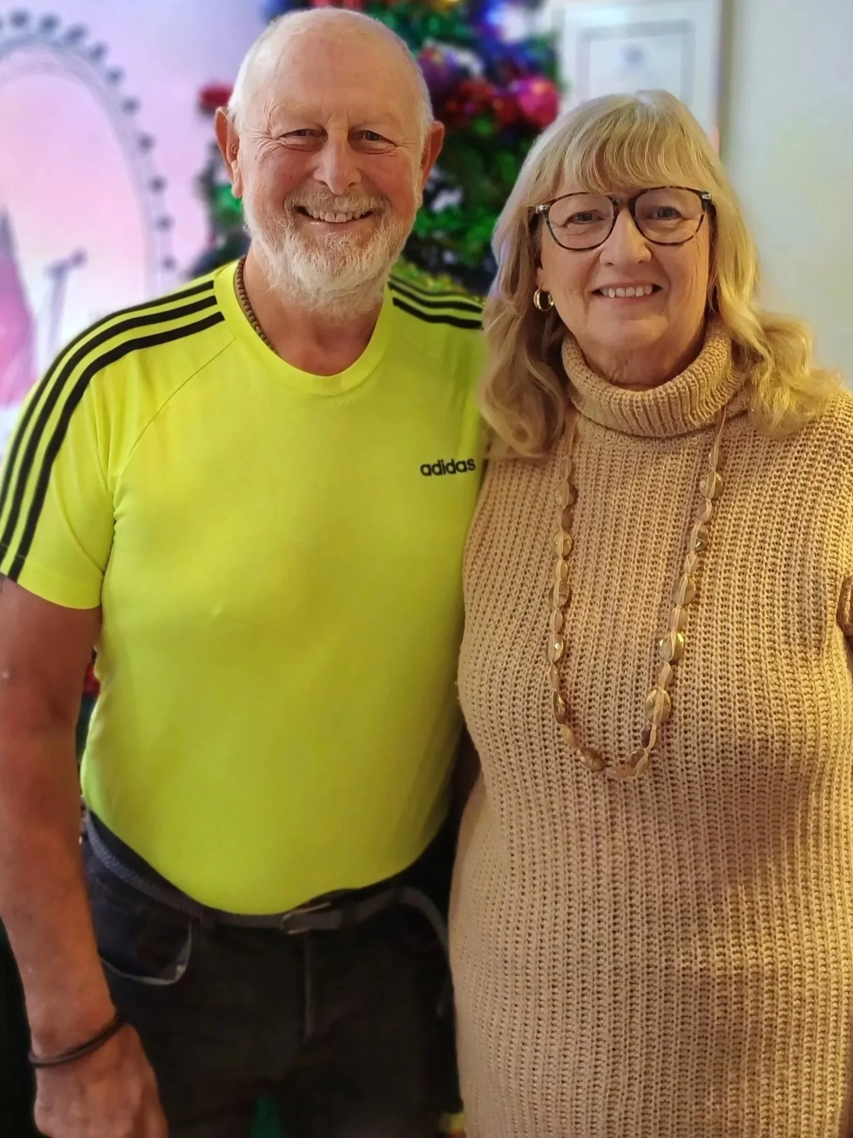 Smiling man with white beard and glasses wearing a bright yellow Adidas shirt, standing next to a woman with blonde hair, glasses, and wearing a beige turtleneck sweater and a long necklace, both indoors with festive decorations in the background.