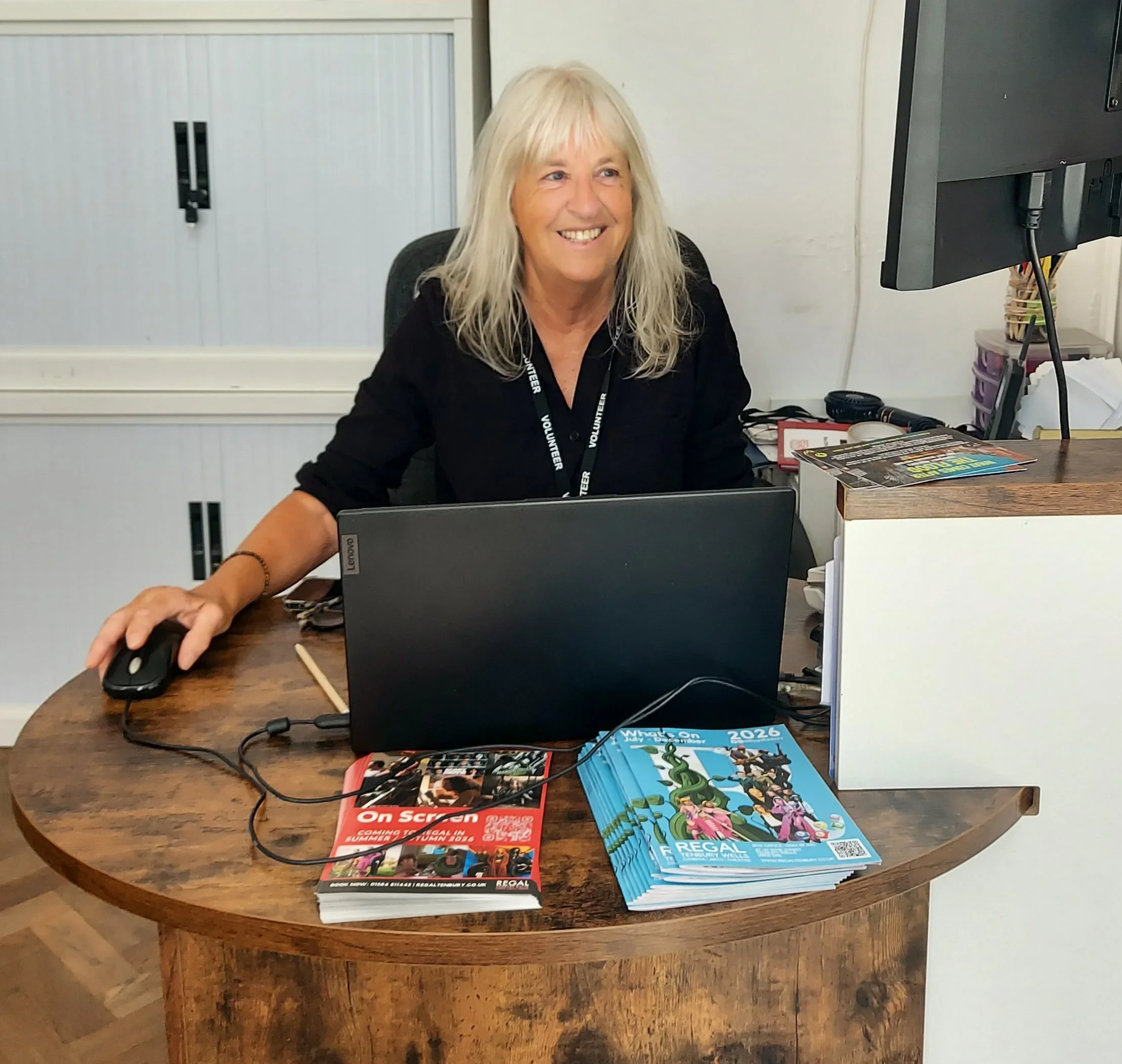 A smiling woman with long, wavy blonde hair sitting at a desk with a computer mouse in her right hand. On the desk, there are brochures, magazines, and a small black device. The background features a locker, a white wall, and a window. The woman wears a black shirt with a volunteer badge hanging around her neck.