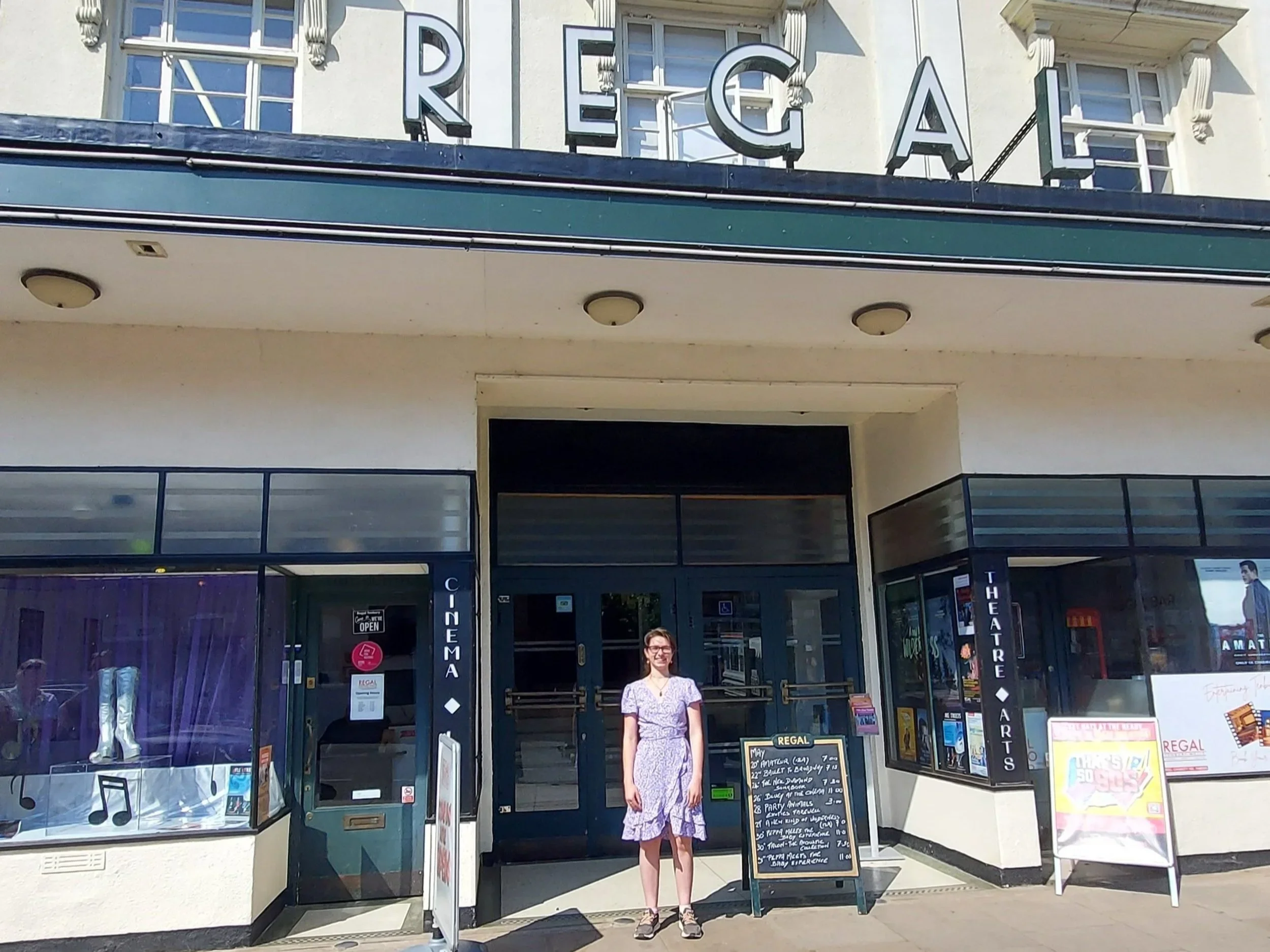 A woman standing in front of a vintage theater entrance with a sign reading 'Regal Theatres.' She is wearing glasses and a purple dress, smiling. The theater's exhibition sign is visible with performances listed, and there are posters and advertisements around the entrance.
