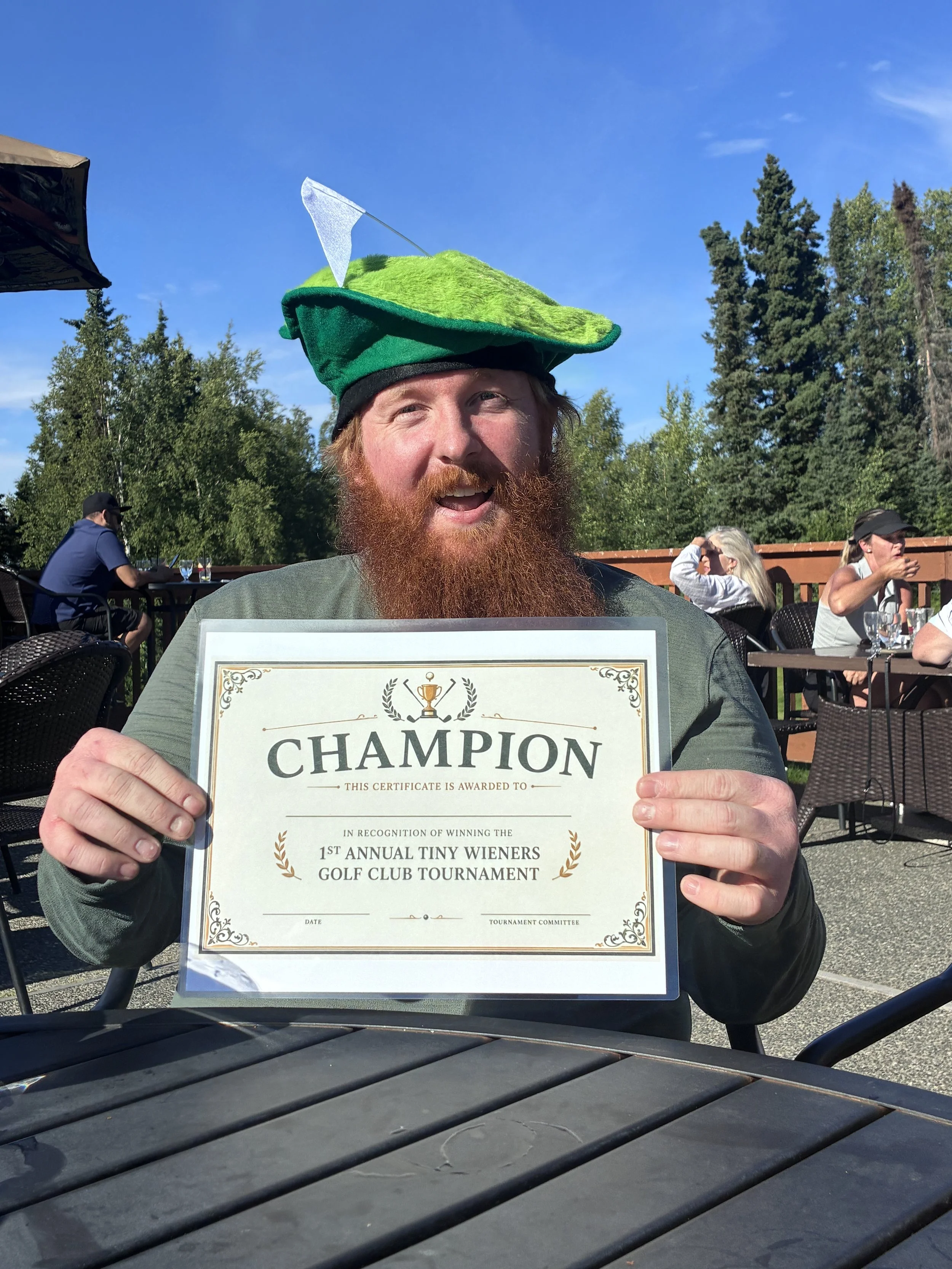 A man with a red beard and a green hat shaped like a golf club head, holding a certificate for winning the 1st Annual Tiny Wieners Golf Club Tournament, outdoors on a sunny day with trees in the background.