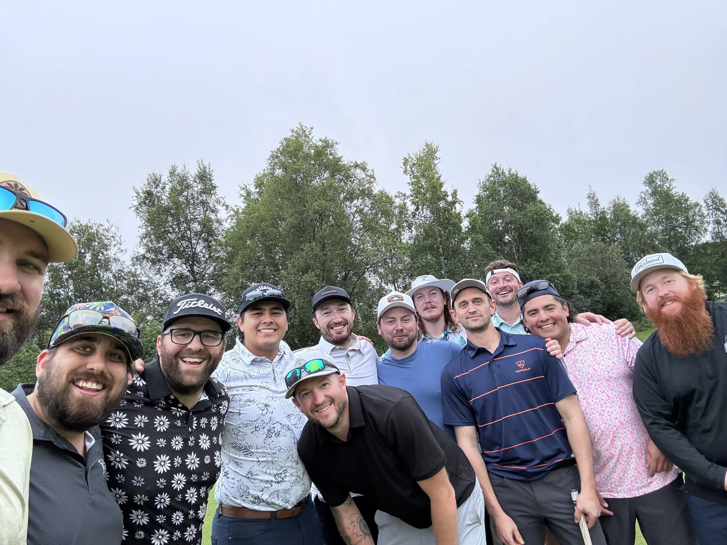 Group of smiling men and women posing outdoors in front of green trees, some wearing hats and sunglasses.