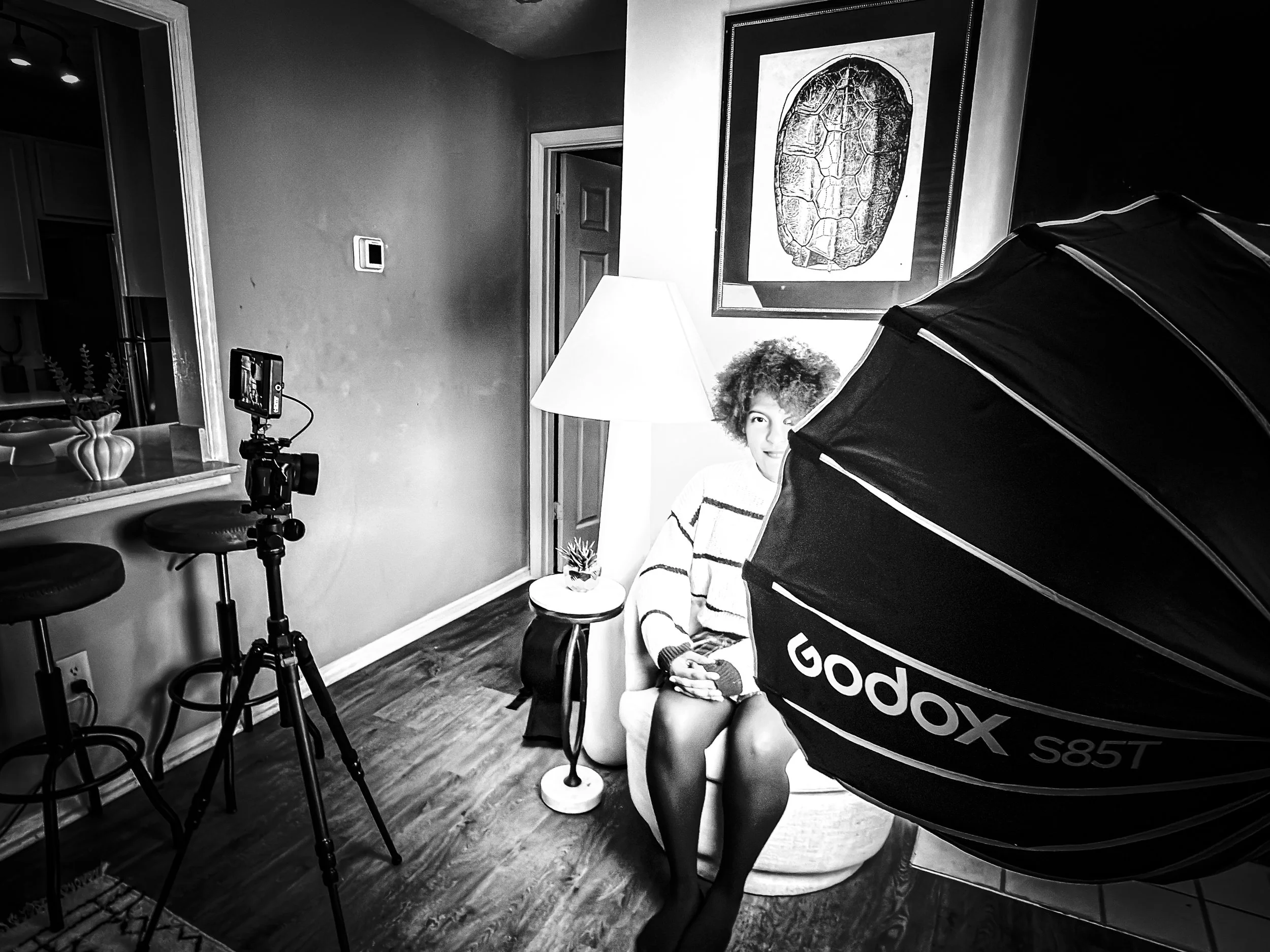 A young woman with curly hair sits in a living room, partially covered by a food photoshoot tent. She appears to be looking at the camera, and there is a large framed picture of a turtle on the wall behind her. Photography equipment including a camera on a tripod and studio lighting are also visible in the room.