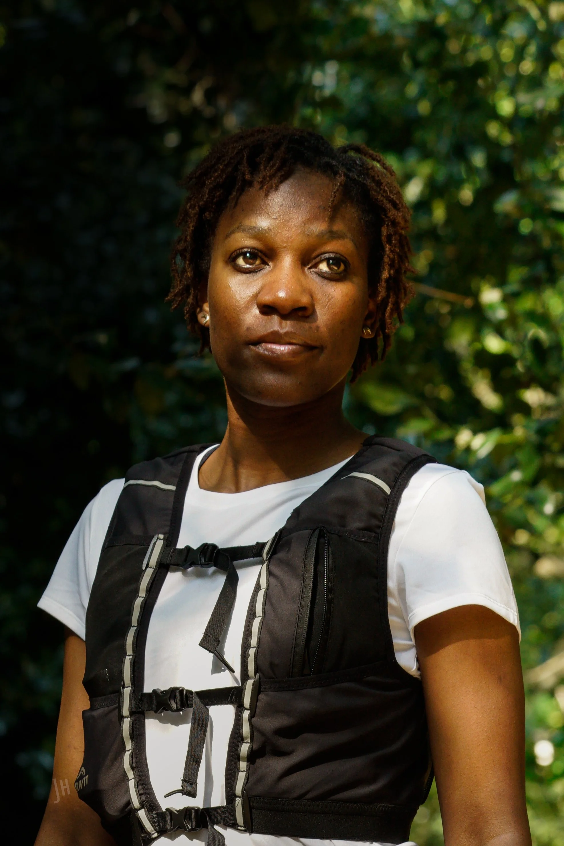 A woman with short dreadlocks and tan skin standing outdoors amidst lush green foliage, wearing a white t-shirt and a black tactical vest.