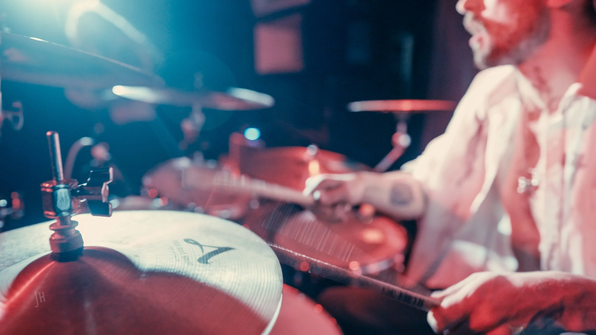 A man with a beard playing an acoustic guitar, wearing a white shirt, in a dimly lit space with red and blue lighting, next to drums.