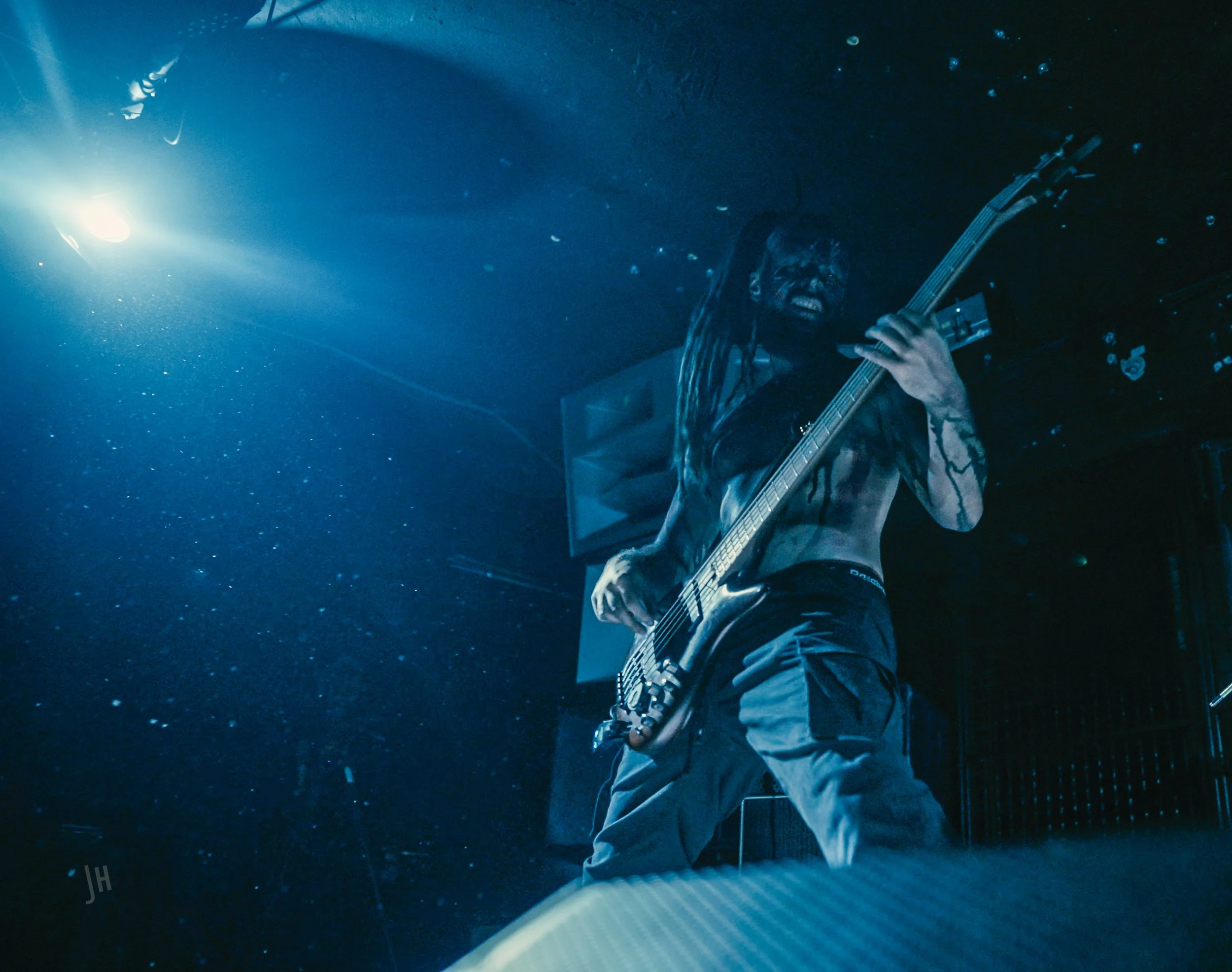 A musician with dreadlocks playing an electric guitar on stage under blue lighting.