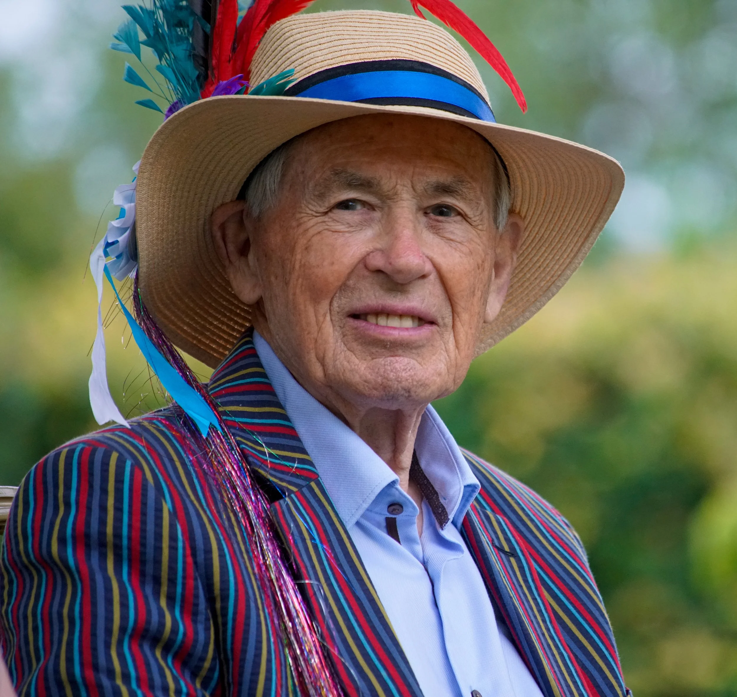 Close-up of an elderly man wearing a colorful striped jacket and a wide-brimmed straw hat decorated with feathers, ribbons, and a blue band, outdoors with a blurred green background.