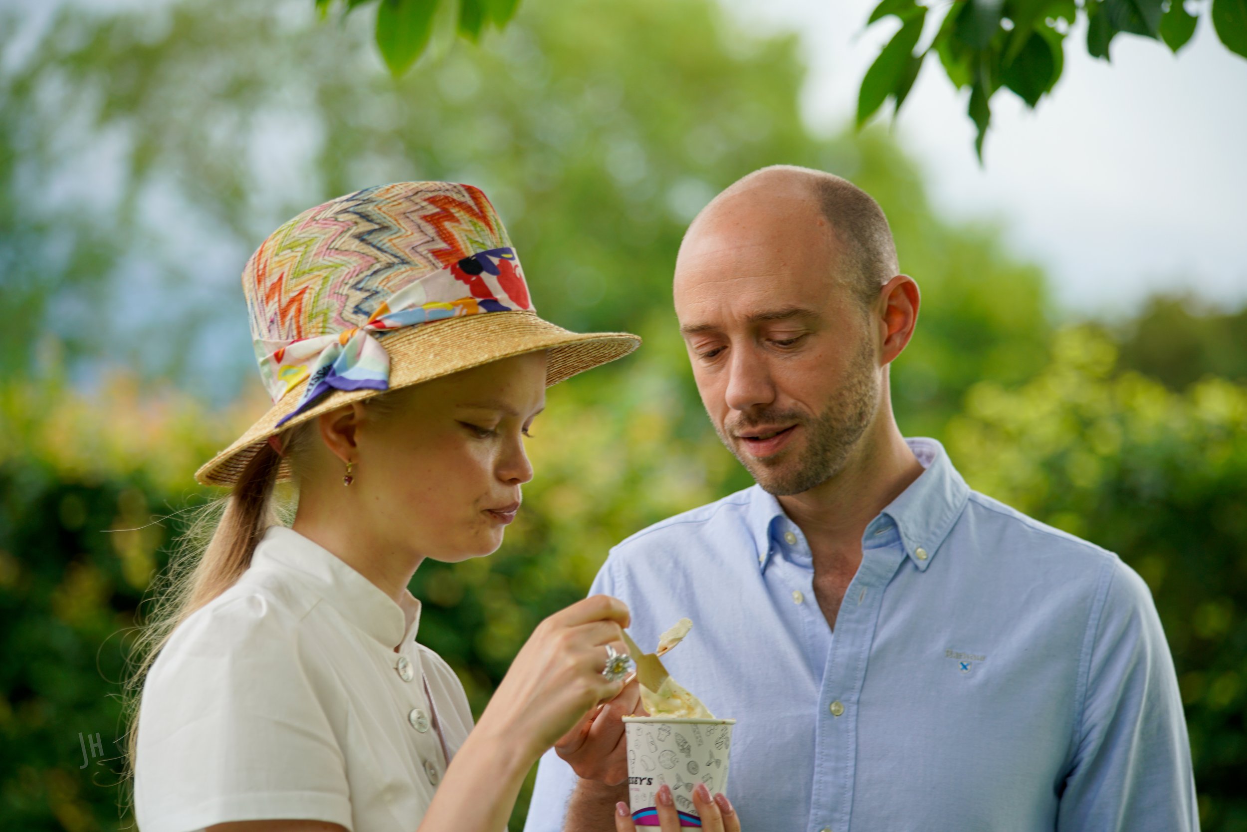 A young girl with a colorful hat eating ice cream while talking to a man in a light blue shirt outdoors under trees.