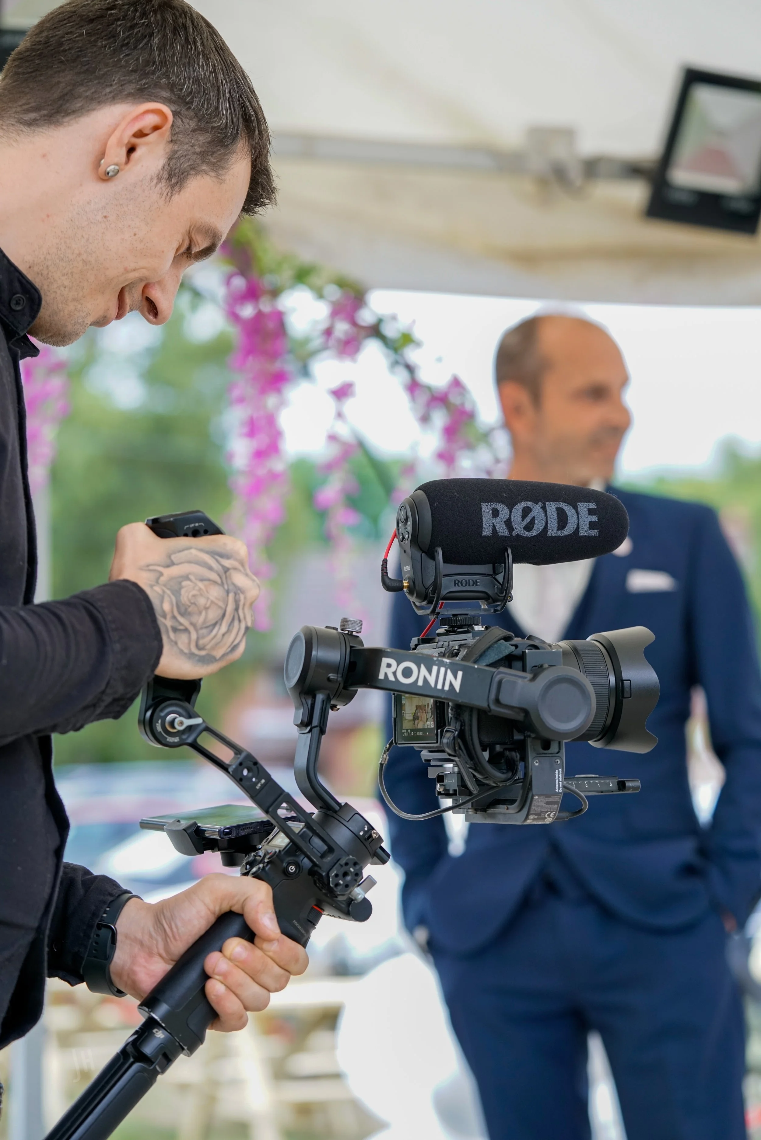 A man with tattoos on his hand operating a professional camera with a stabilizer and a Rode microphone mounted on it. Another man in a suit is blurred in the background, with pink flowers and greenery around.