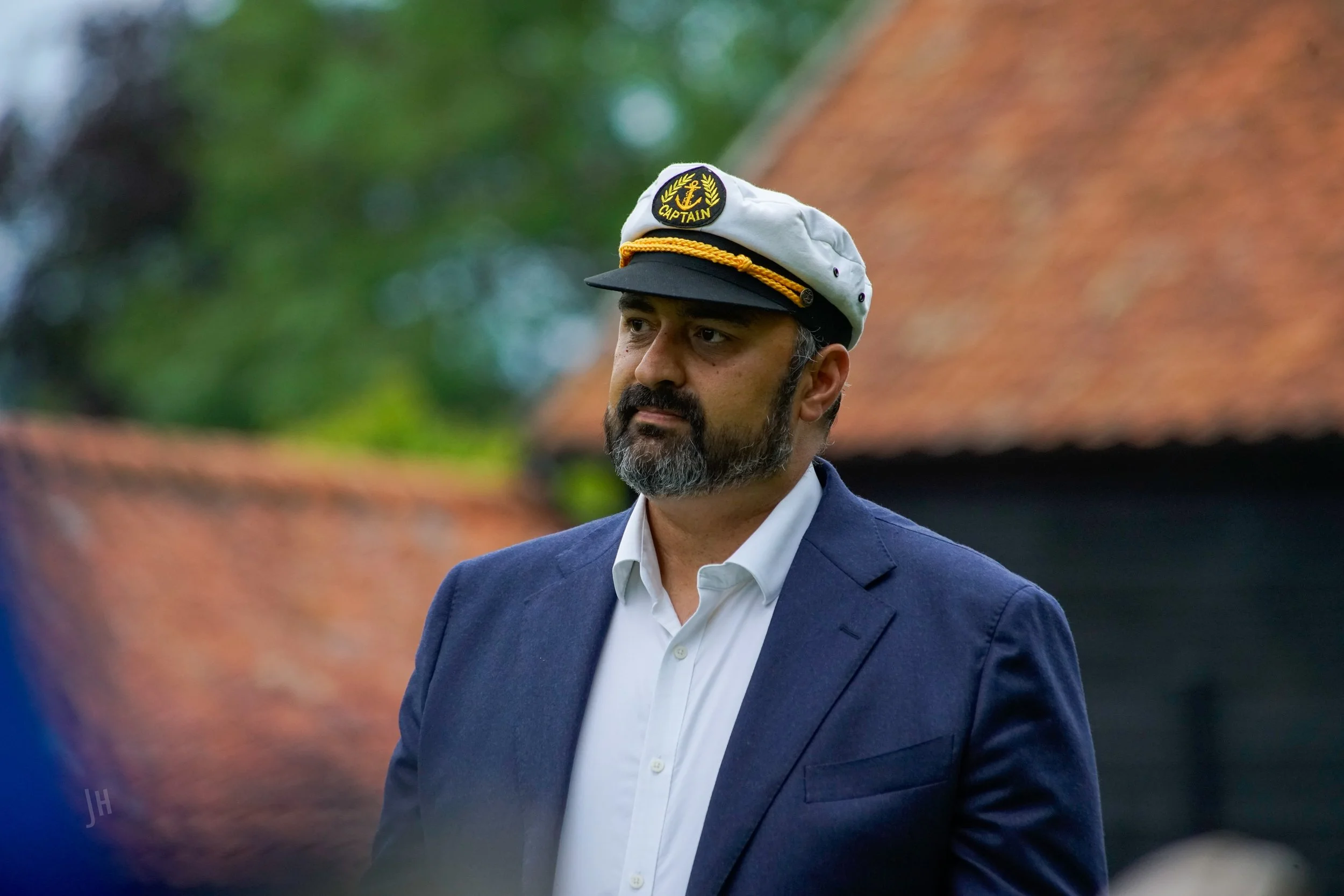 A man with a beard wearing a captain's hat and a navy blue suit jacket over a white shirt, standing outdoors with a blurred background of trees and a building with a red roof.