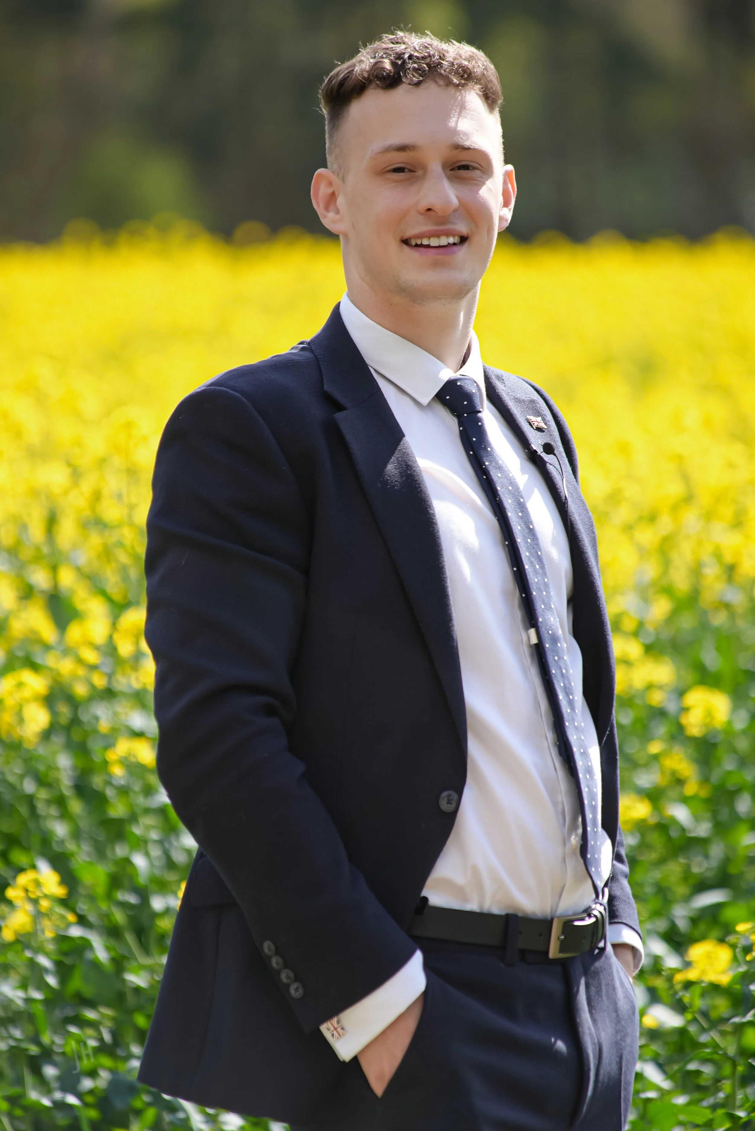 Young man in a suit standing outdoors in a field of yellow flowers with greenery and trees in the background, smiling at the camera.
