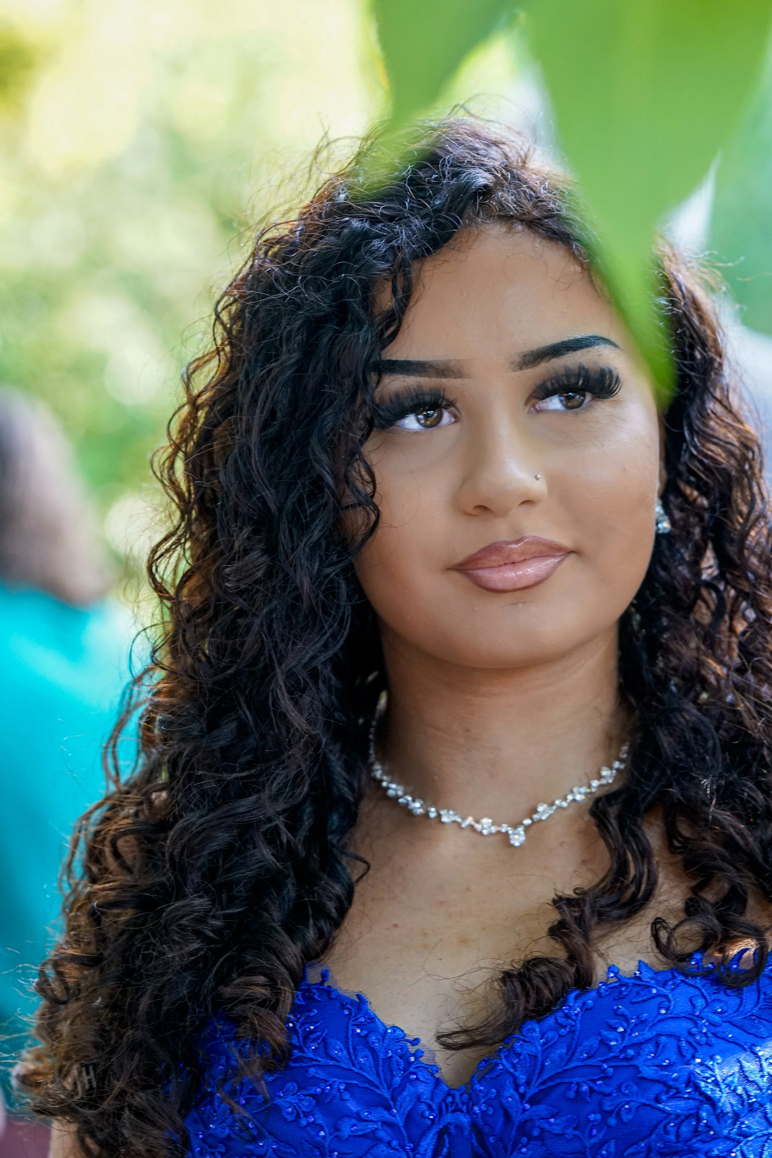 A woman with curly black hair, brown eyes, and a nose piercing, wearing a blue dress with embroidery, a pearl necklace, and earrings, outdoors with green background.