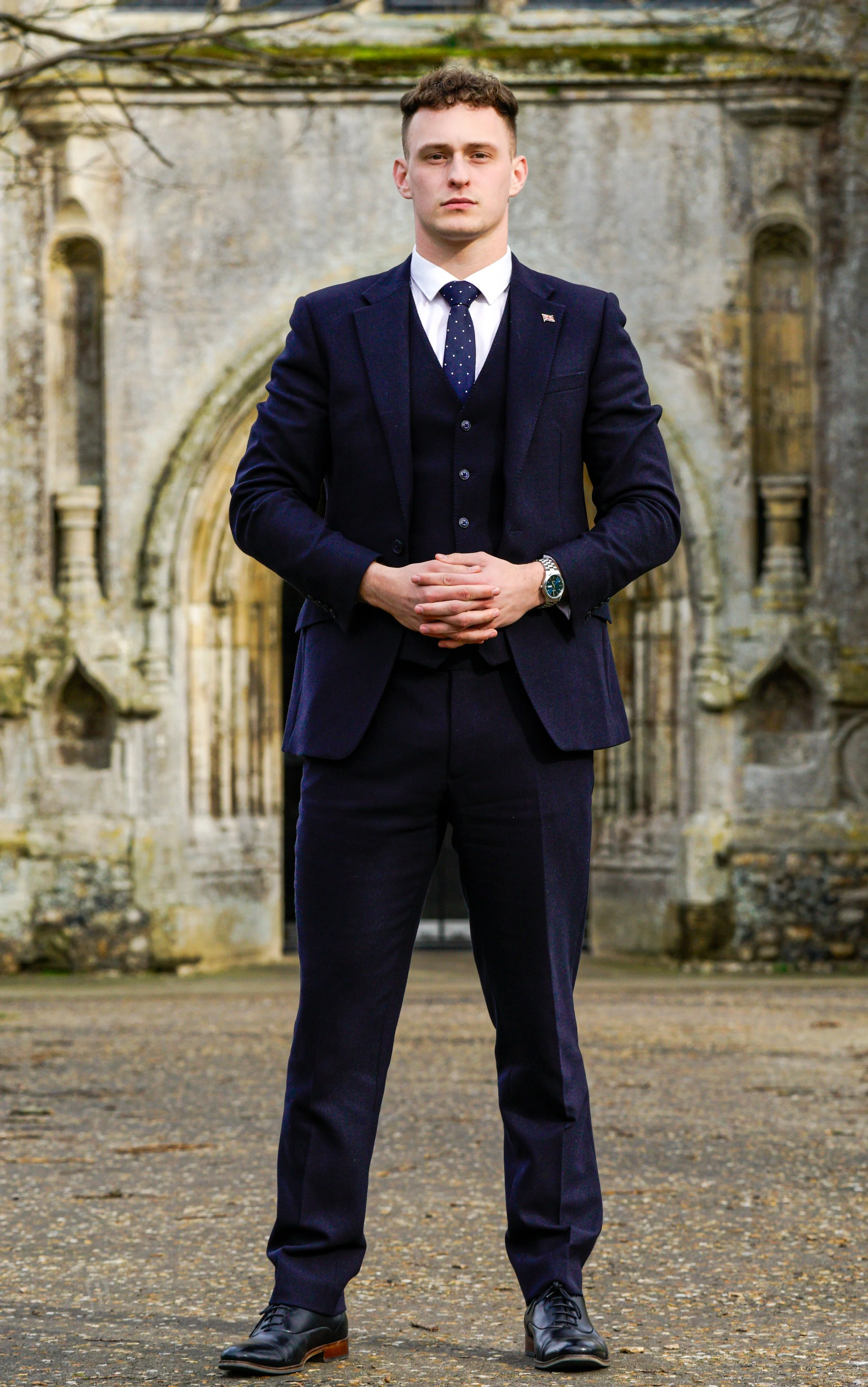 A young man in a dark suit, white shirt, and navy tie with white dots stands with hands clasped in front of him in front of an old stone building with an arched doorway and weathered walls.
