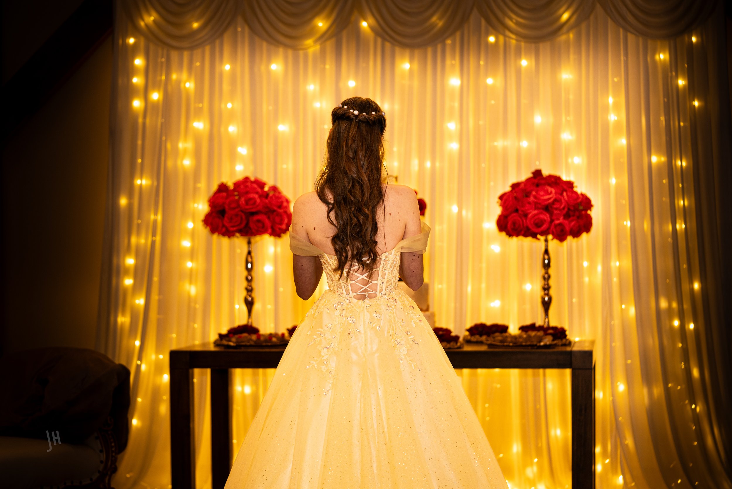 Back of a woman in a yellow gown with lace-up back, standing in front of a decorated backdrop with gold curtains, string lights, and red flower arrangements.