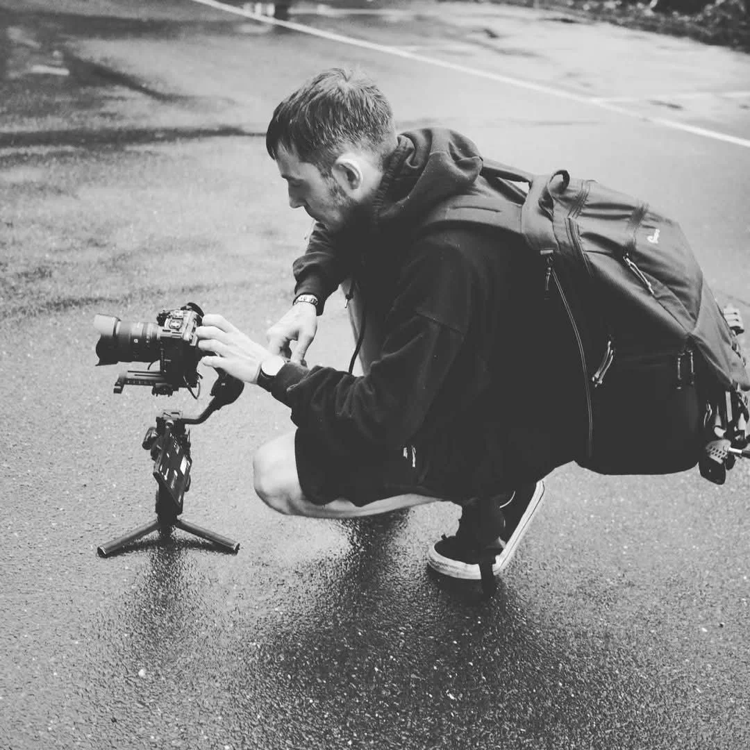 A man with a backpack kneeling on a wet pavement, adjusting a camera mounted on a small tripod.