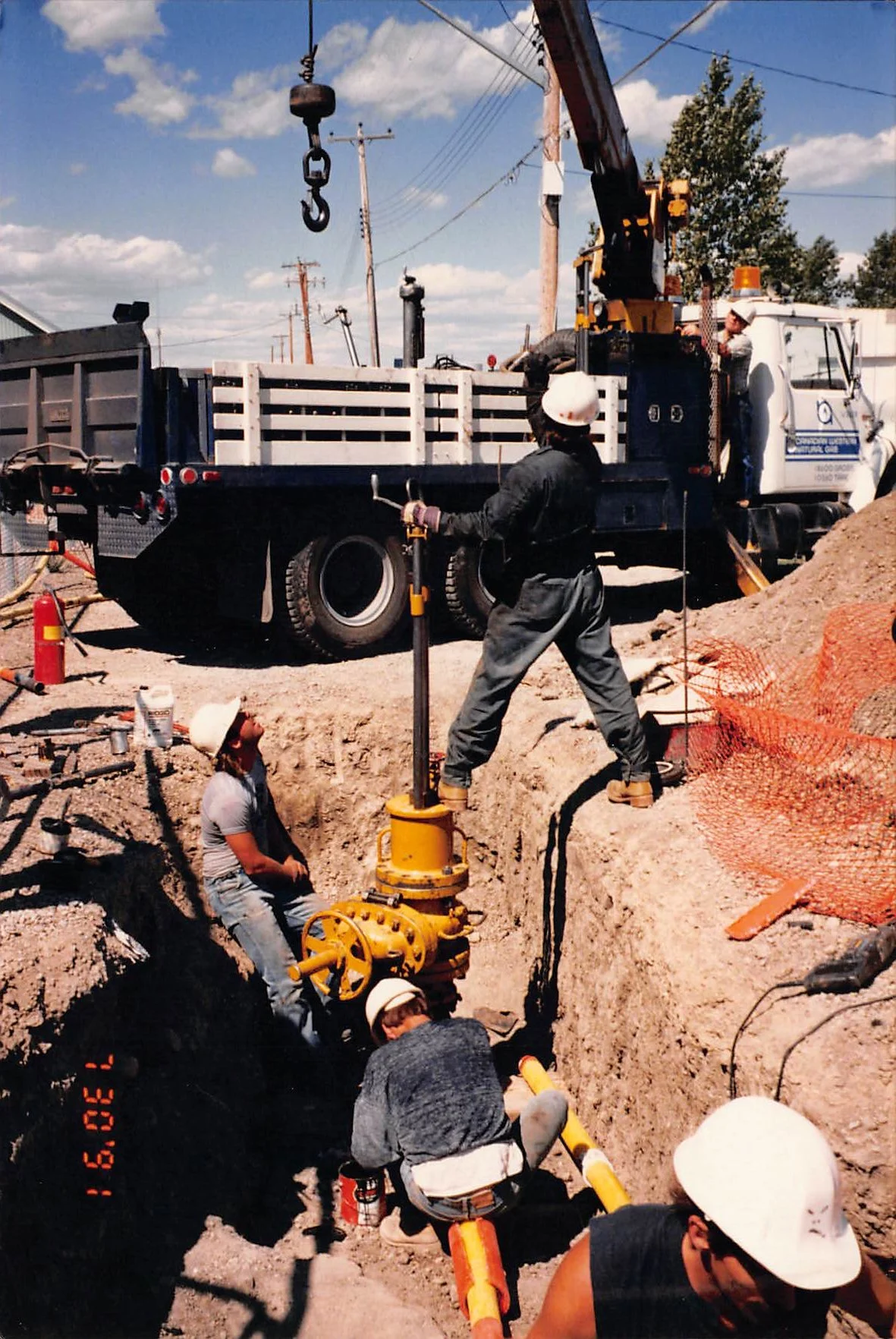 Workers at a construction site installing underground utility pipes, with a large truck and crane in the background, under a sunny sky.