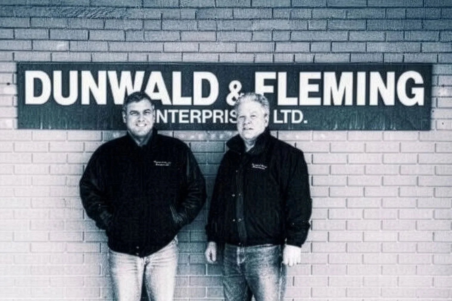 Two men standing in front of a sign that reads 'DUNWALD & FLEMING ENTERPRISES LTD.' on a brick wall. Both are wearing black jackets.