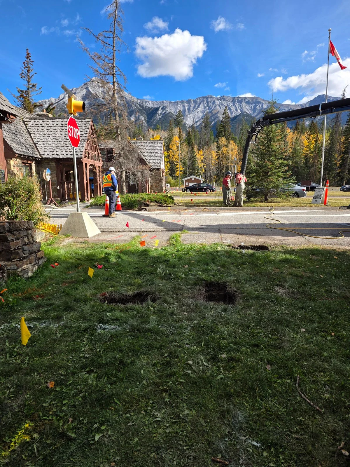Workers operating equipment and digging holes on a road beside a building with a stop sign, surrounded by trees and mountains under a partly cloudy sky.