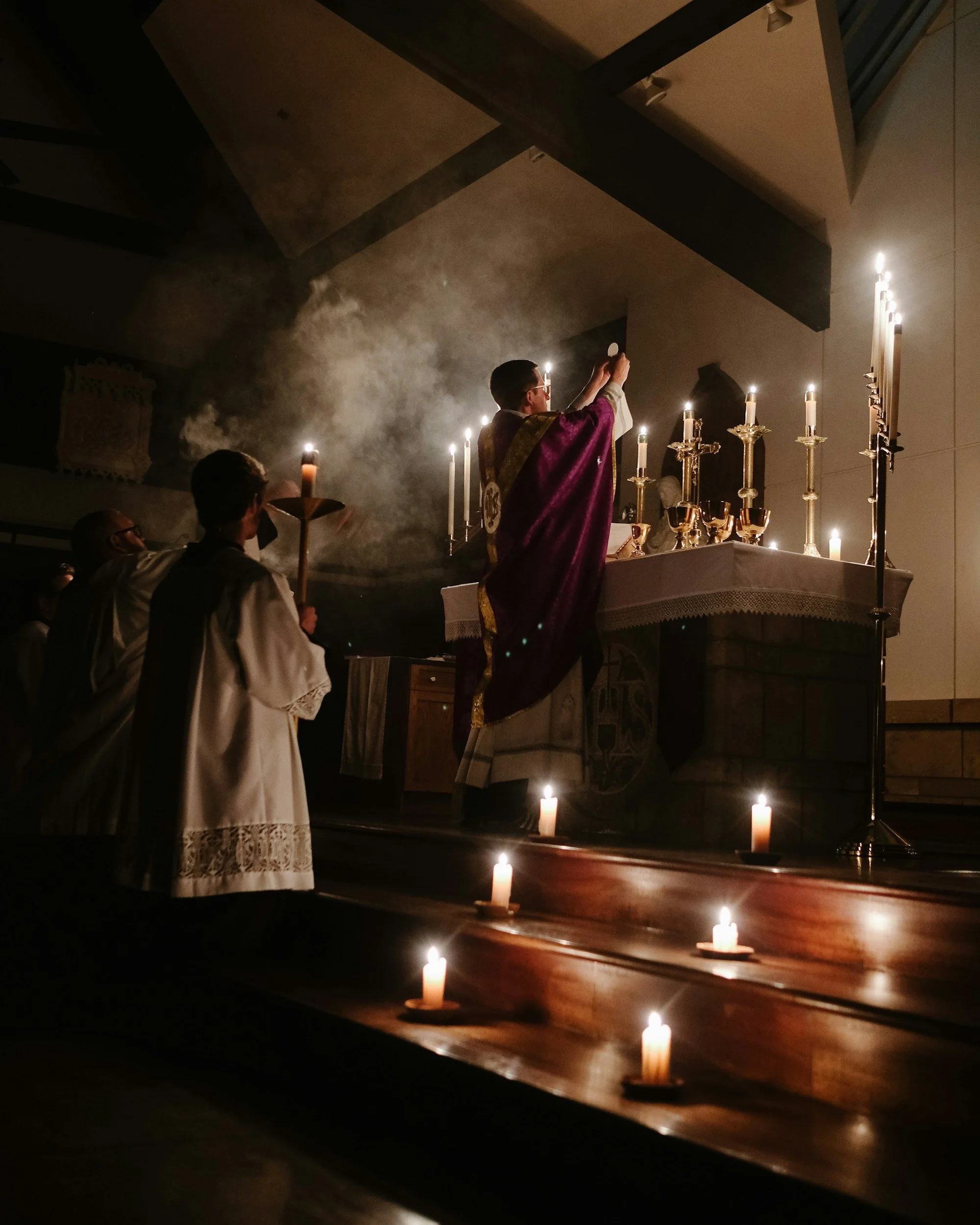 Clergy member in liturgical vestments leading a religious service at an altar decorated with lit candles, with other clergy or participants in the background, in a dimly lit church.