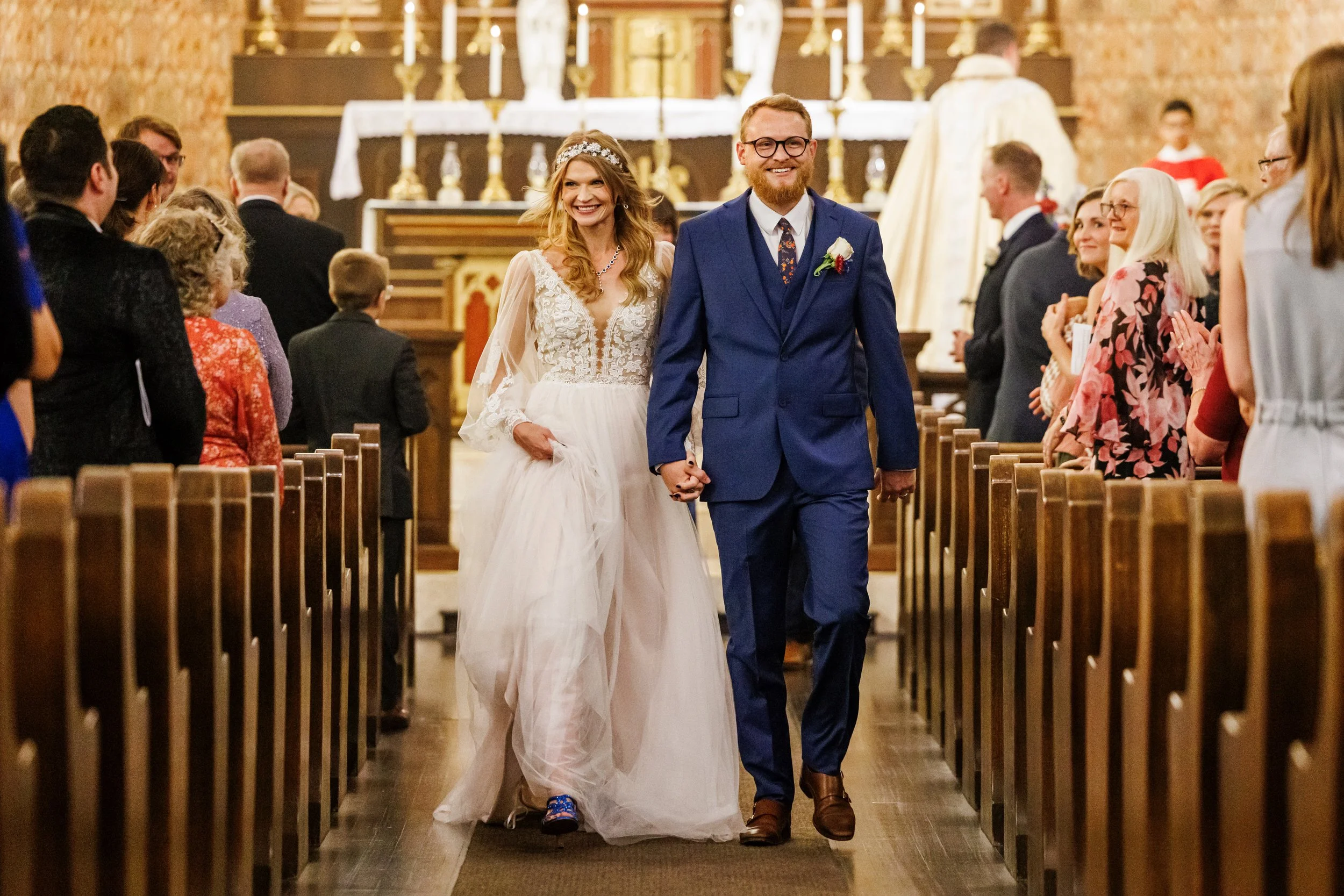 A smiling couple walking down the aisle of a church during their wedding, holding hands. The bride is wearing a white wedding dress and a tiara, while the groom is dressed in a blue suit with glasses. Guests are standing on either side, clapping and smiling.