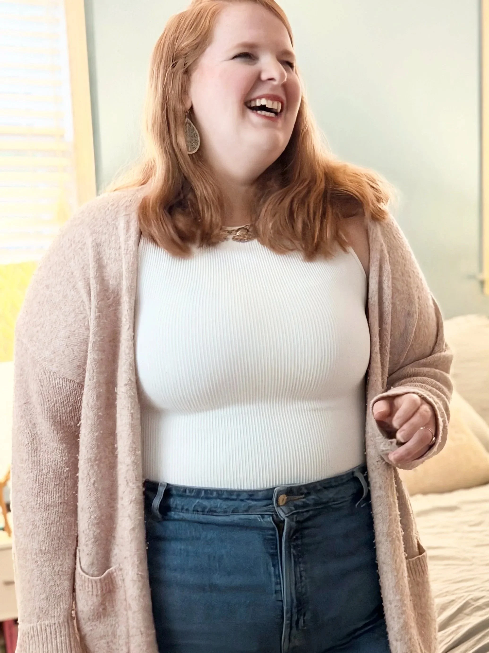 Close-up of a woman with red hair wearing a white top, blue jeans, and a beige cardigan, standing indoors in front of a bed with a light-colored wall and window blinds.