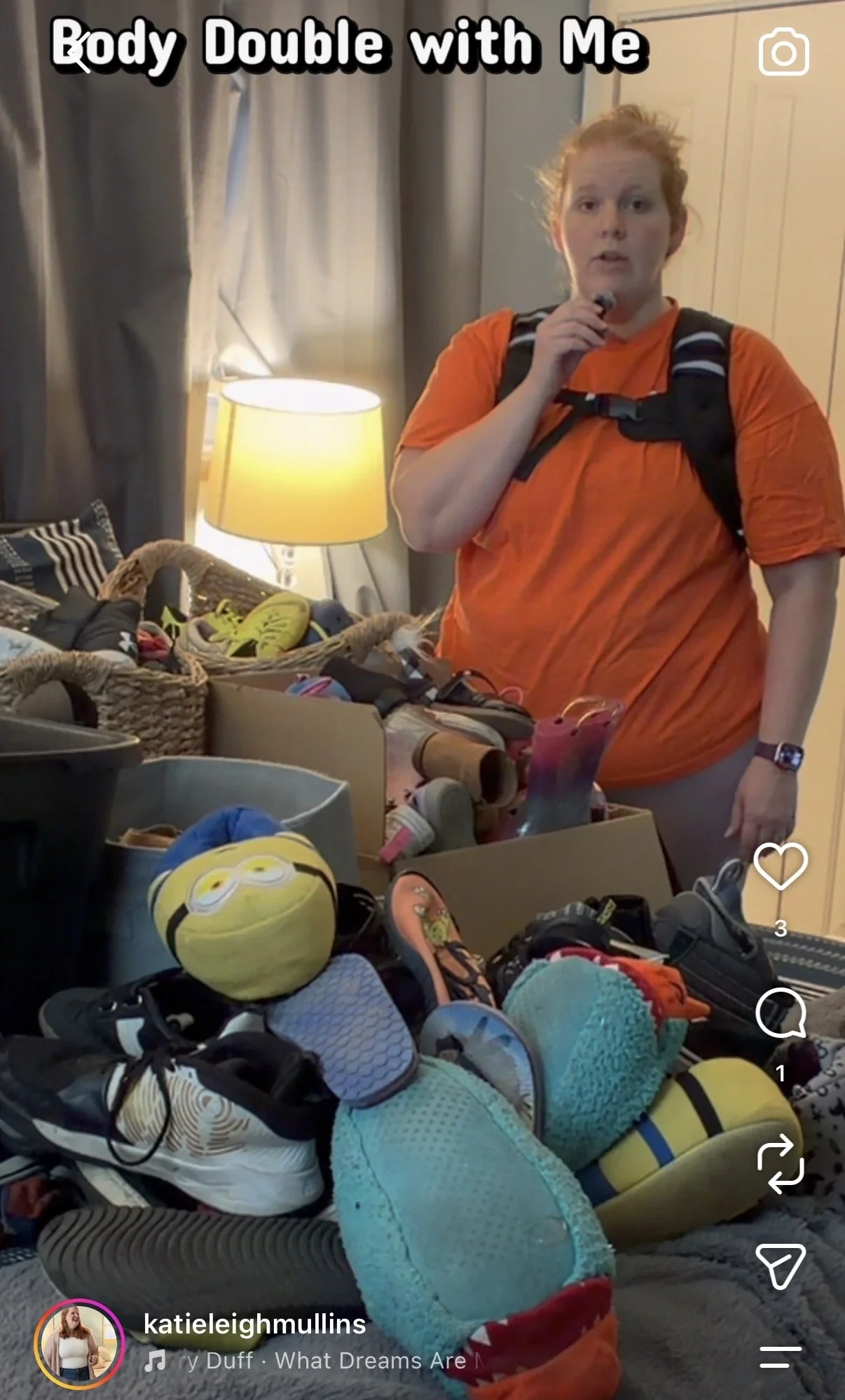 A woman wearing an orange shirt and a backpack standing in a room with many shoes, toys, and items on a table, illuminated by a lamp, with curtains in the background and the text 'Body Double with Me' at the top.