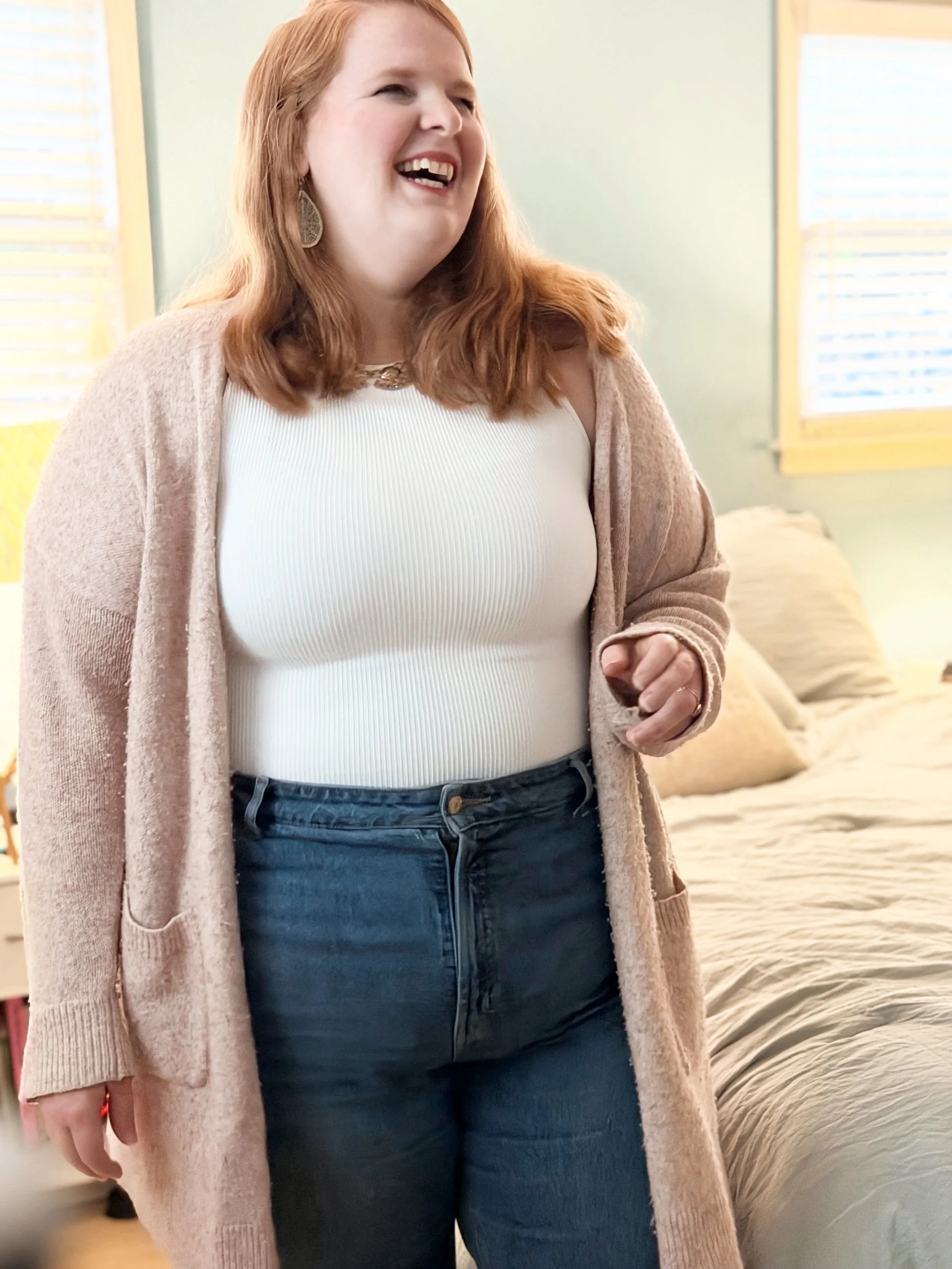 A woman with red hair, wearing a white top, dark jeans, and a beige cardigan, smiling inside a room with light-colored walls and windows.